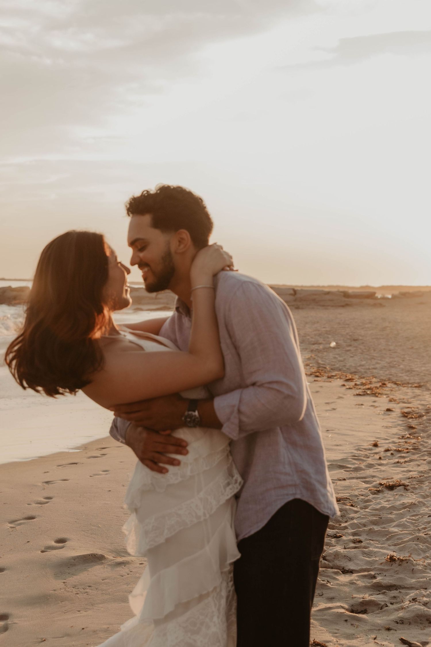 Couple embracing romantically on a sandy beach at golden sunset, bride in white dress and groom in linen shirt.
