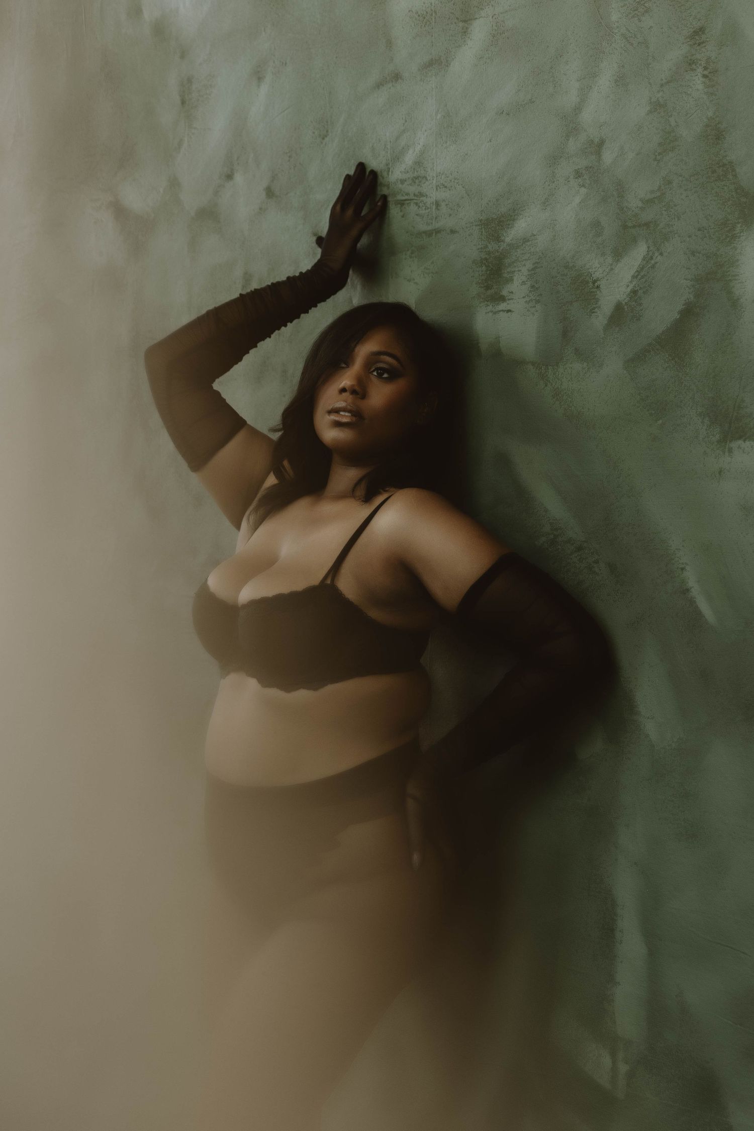 A plus-size Black woman in black lingerie and gloves poses gracefully against a textured green wall in moody lighting.
