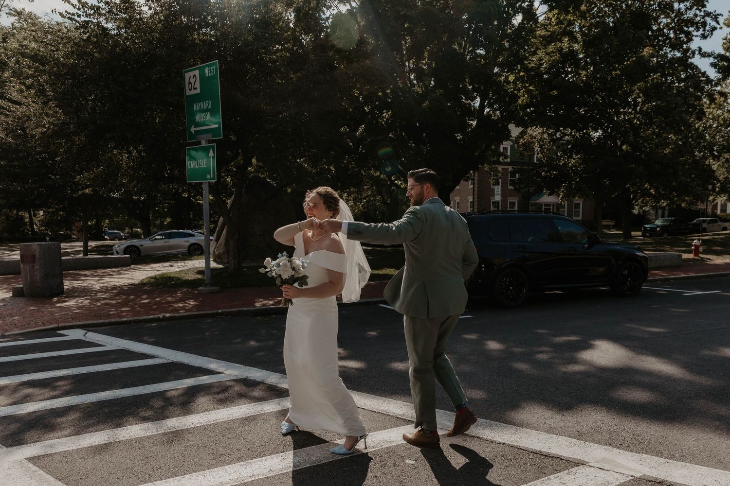 Bride and groom crossing a sunny street in wedding attire, bride holding white bouquet, groom in green suit.