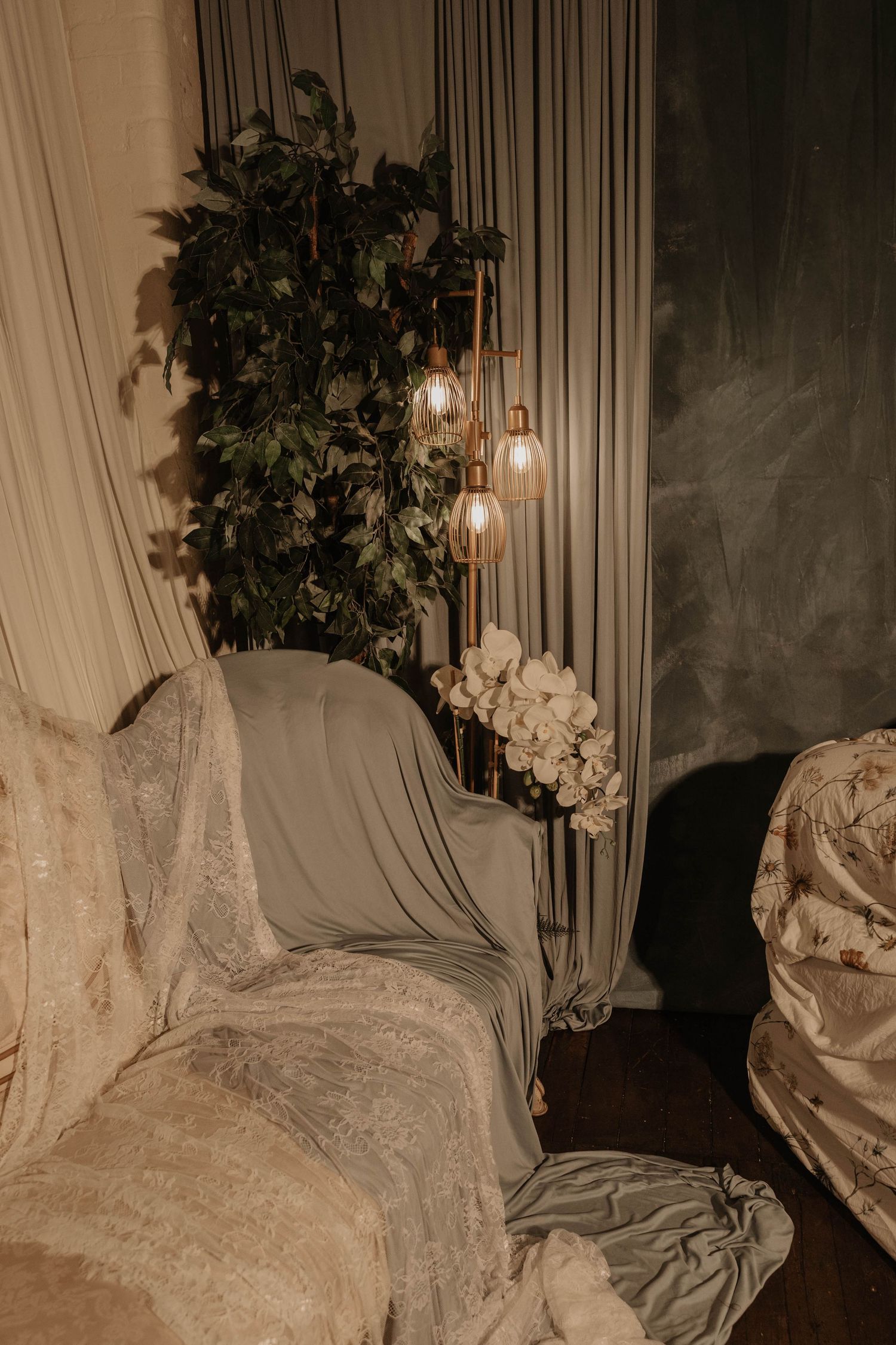 Elegant wedding lounge corner with draped fabric, lush greenery, Edison bulb lighting, and white floral arrangement.