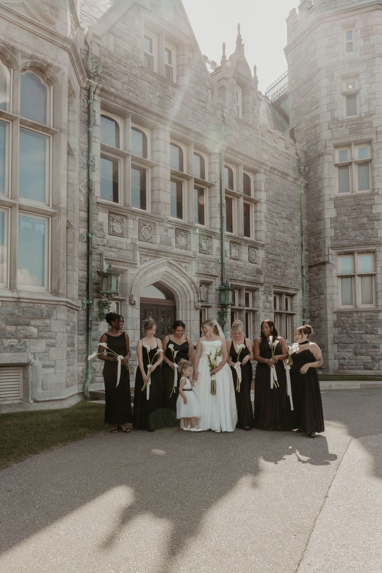 Bride and bridesmaids in black gowns pose before a grand stone castle wedding venue.