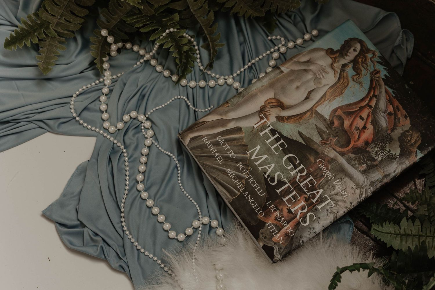 Book 'The Great Masters' with pearl necklace and blue fabric on white surface with greenery.