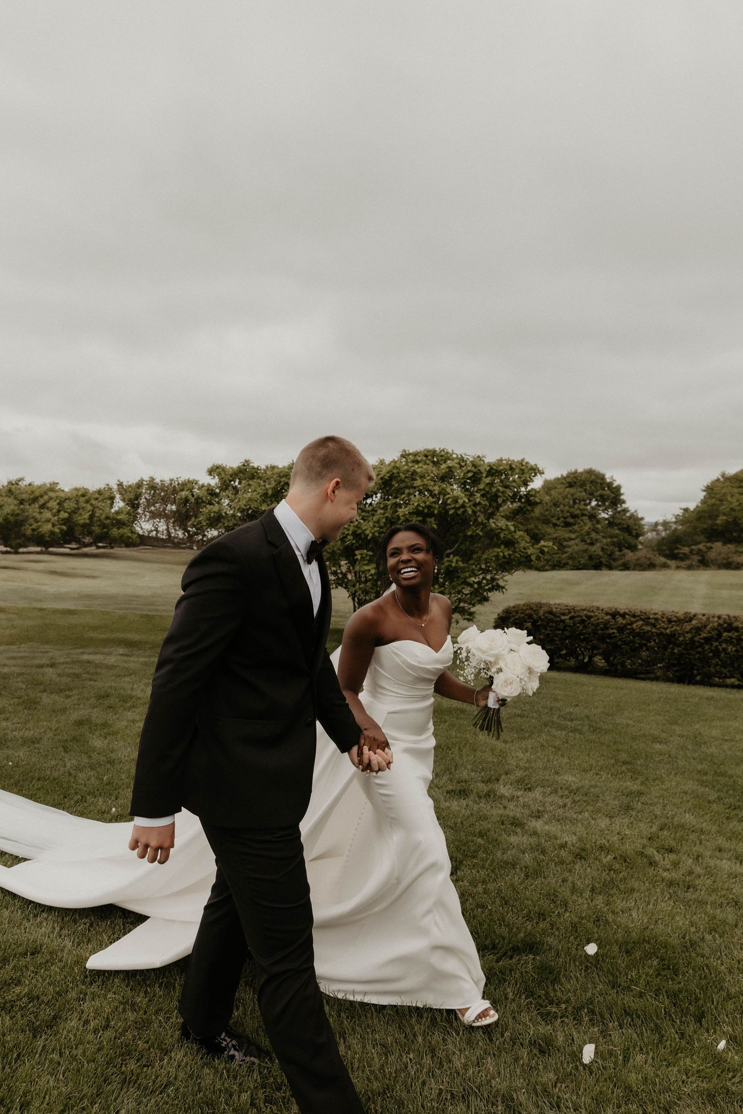 Joyful interracial bride and groom walking hand in hand on green lawn, bride in strapless gown holding white bouquet.