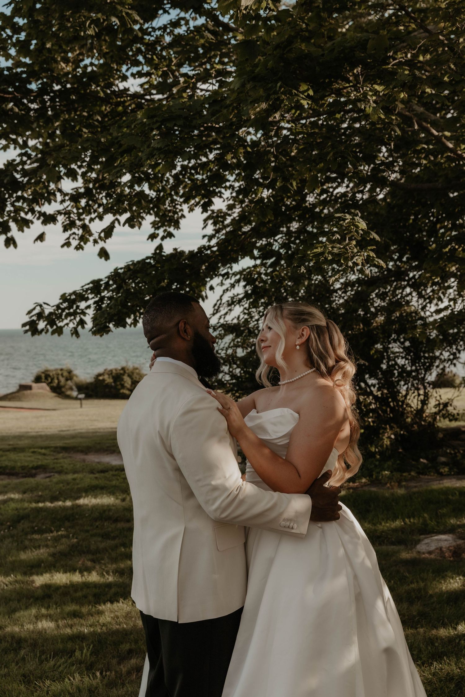 A bride and groom share a romantic moment under a tree near the water, smiling at each other on their wedding day.