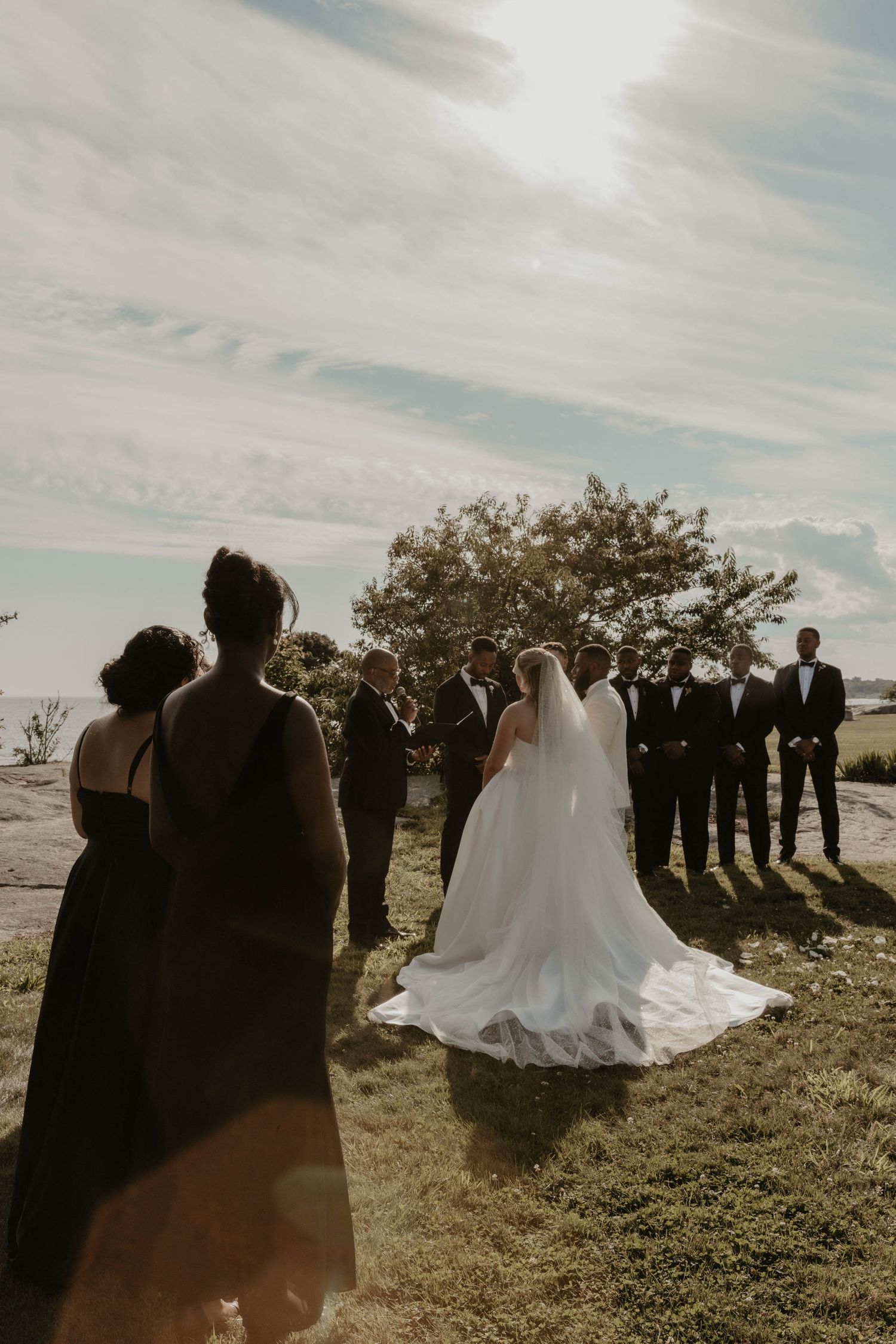 Outdoor wedding ceremony with bride in white gown and veil, groom, groomsmen in black tuxedos, and bridesmaids on a grassy field.