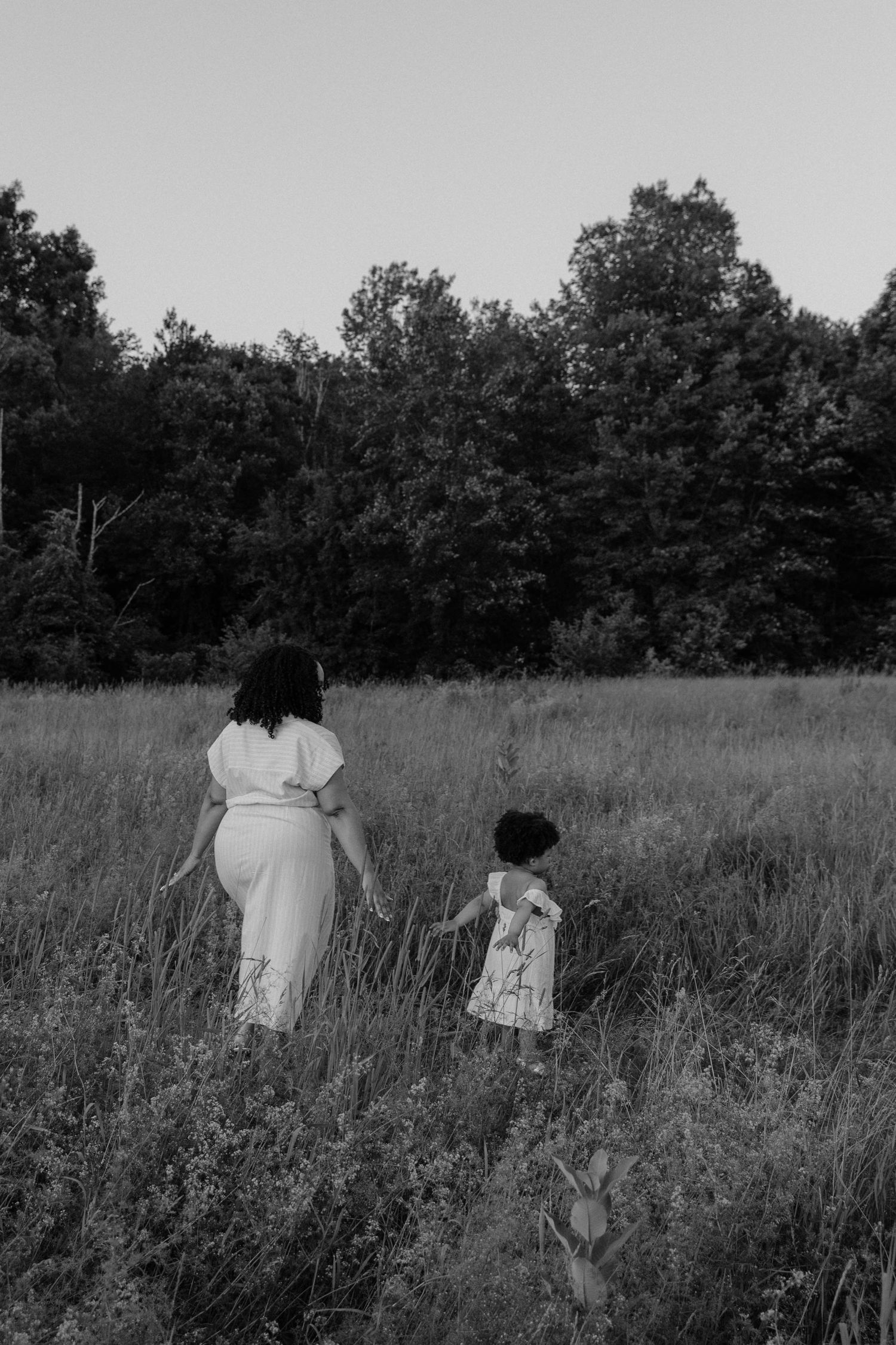 Black and white photo of a mother and young daughter walking hand in hand through a grassy meadow near trees.