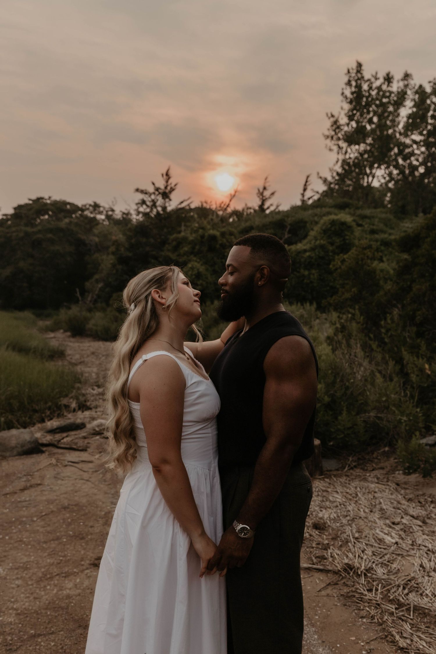 Interracial couple holding hands at sunset, woman in white dress and man in black tank top, surrounded by lush greenery.