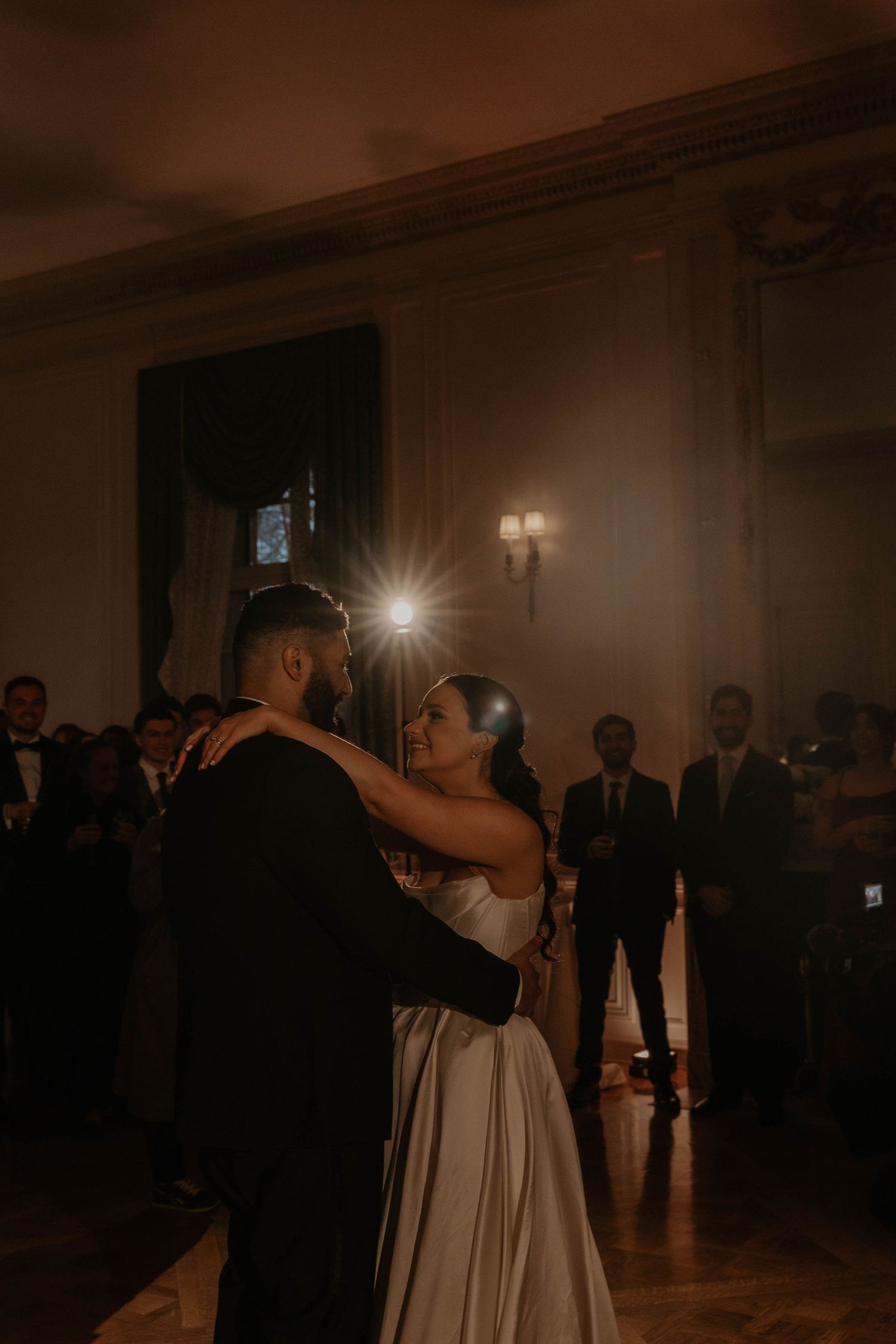 Bride and groom share their first dance in an elegant dimly lit ballroom surrounded by wedding guests.