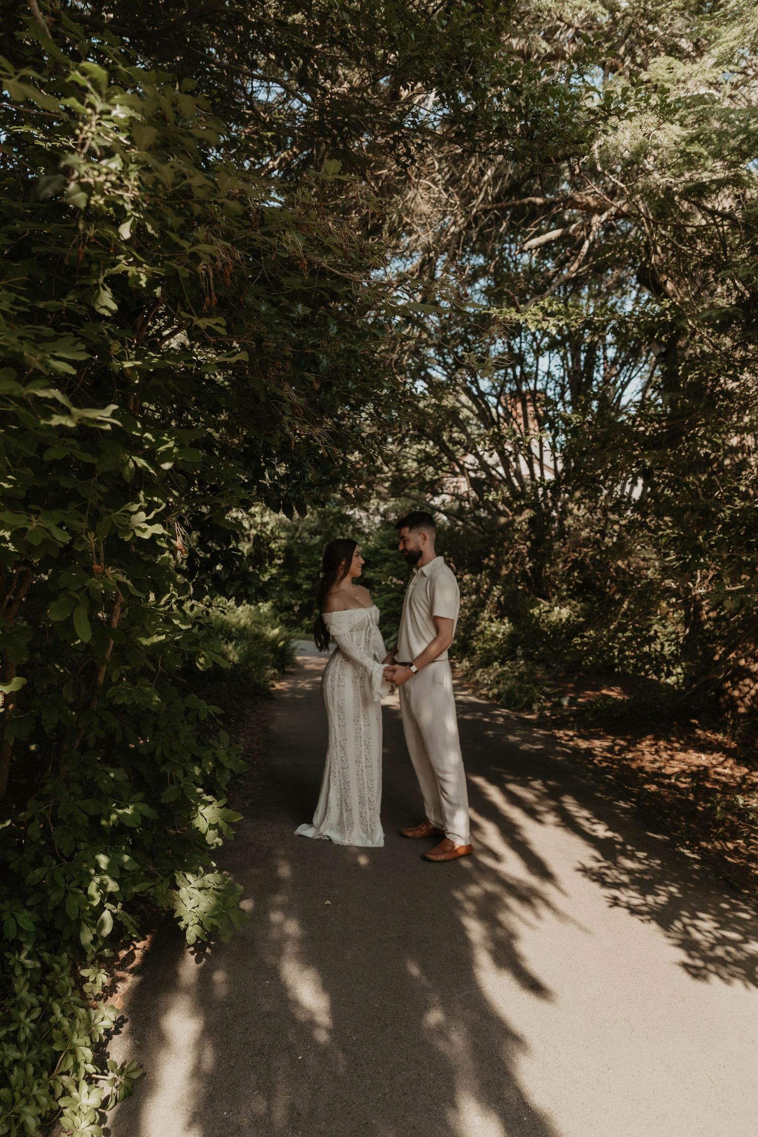 Couple holding hands on a sunlit garden path surrounded by lush green trees in wedding attire.