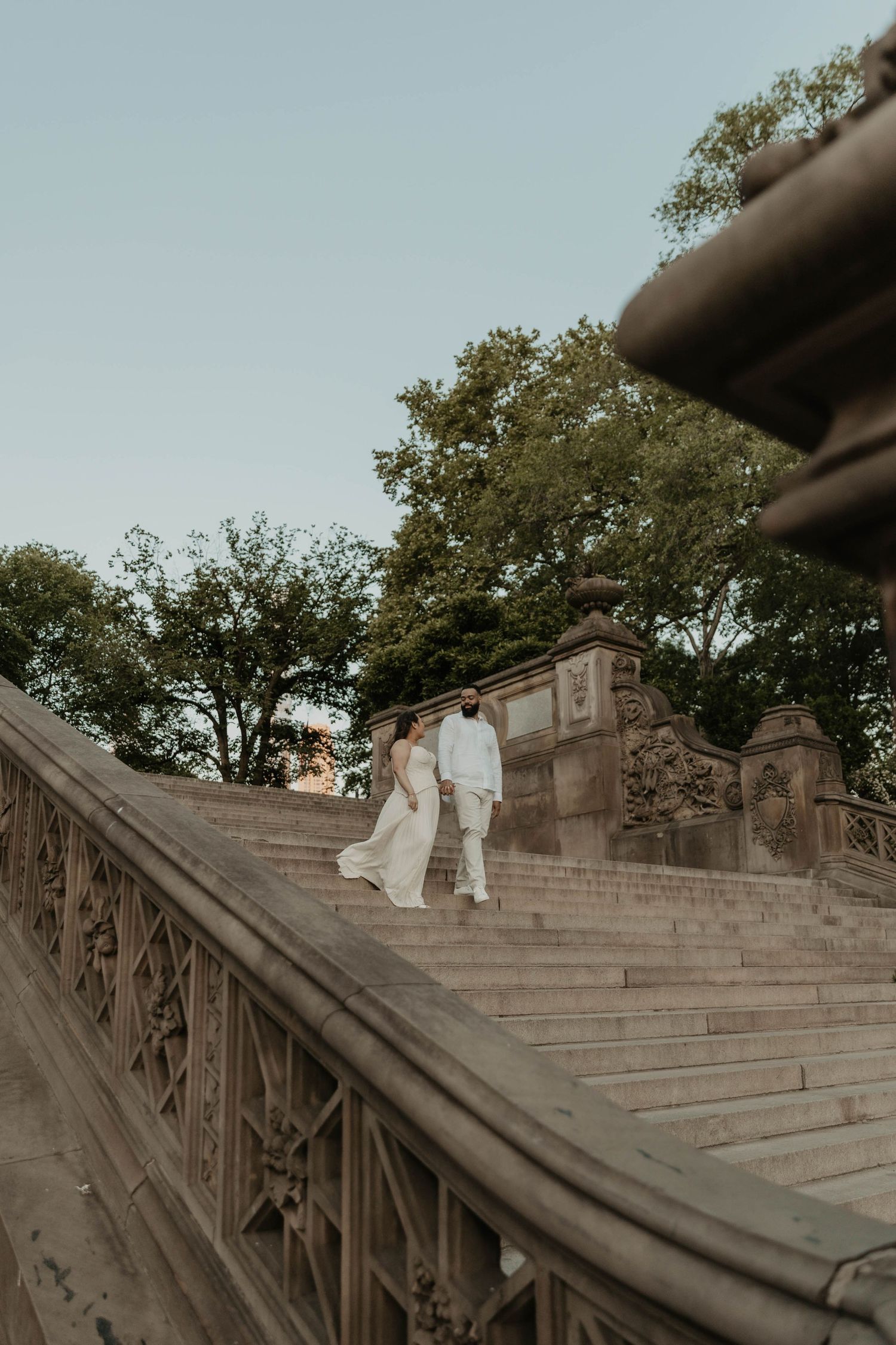 A couple in white wedding attire walks up ornate stone steps at Bethesda Terrace in Central Park at dusk.
