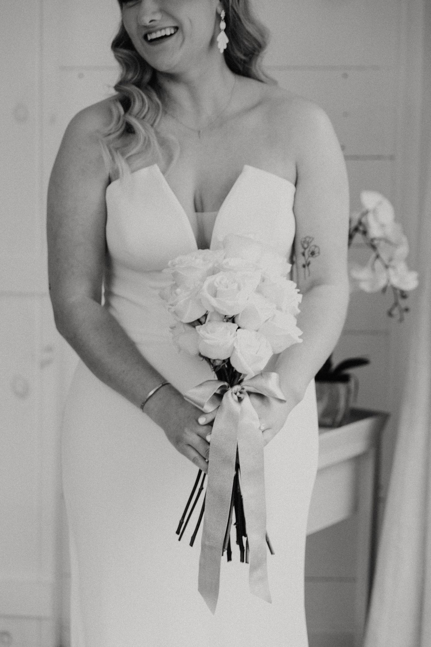 Black and white photo of a smiling bride in a strapless gown holding a white rose bouquet tied with a long satin ribbon.