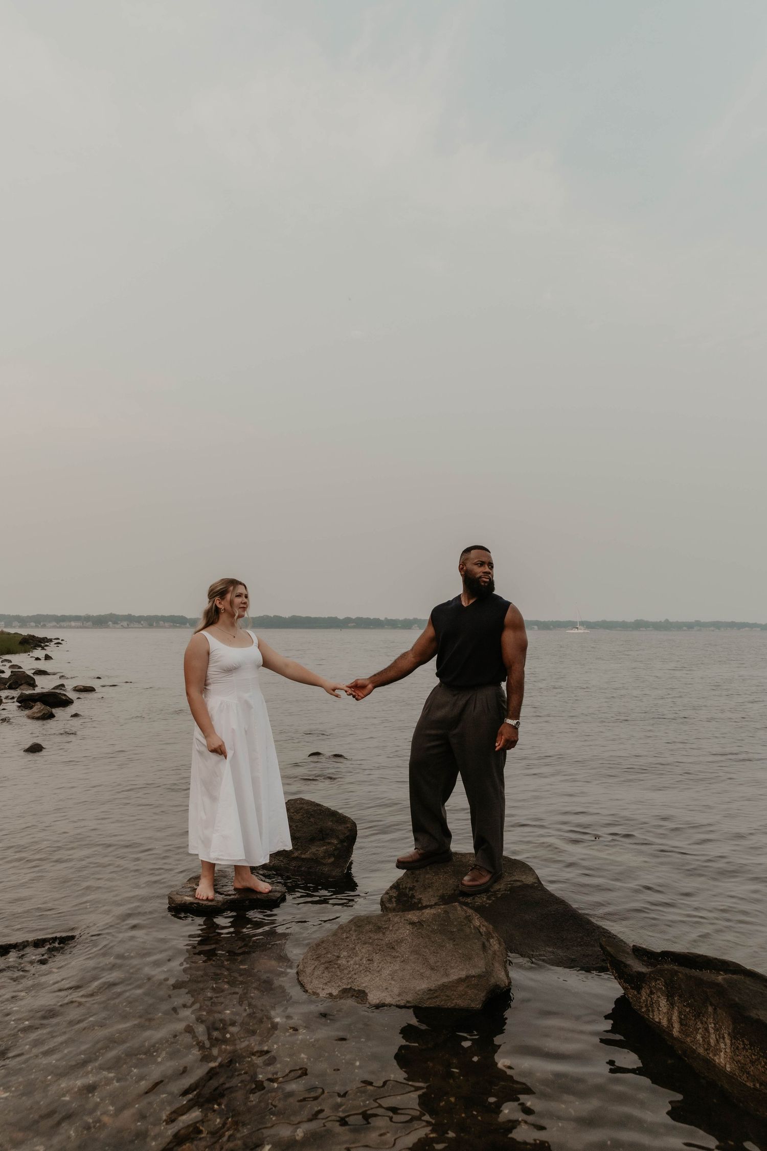 Couple holding hands on rocky shoreline, woman in white dress and man in dark outfit, standing by calm water under overcast sky.