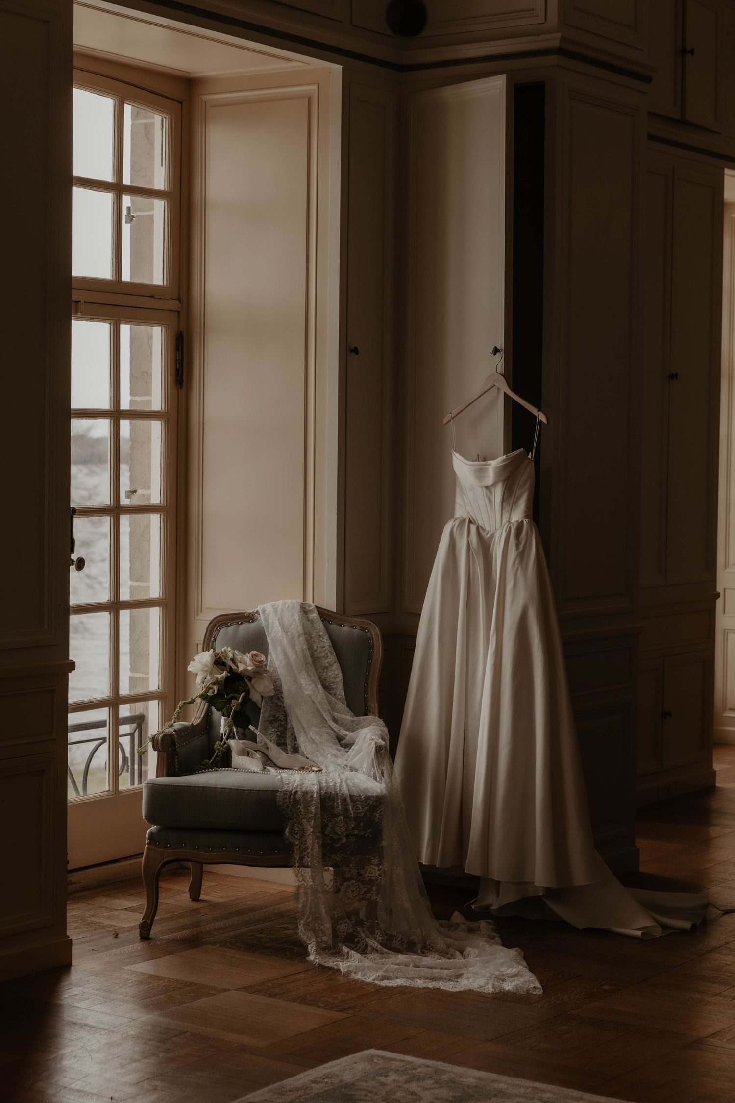 Elegant wedding dress hanging in a sunlit vintage room with a lace veil draped over an antique chair beside a floral bouquet.
