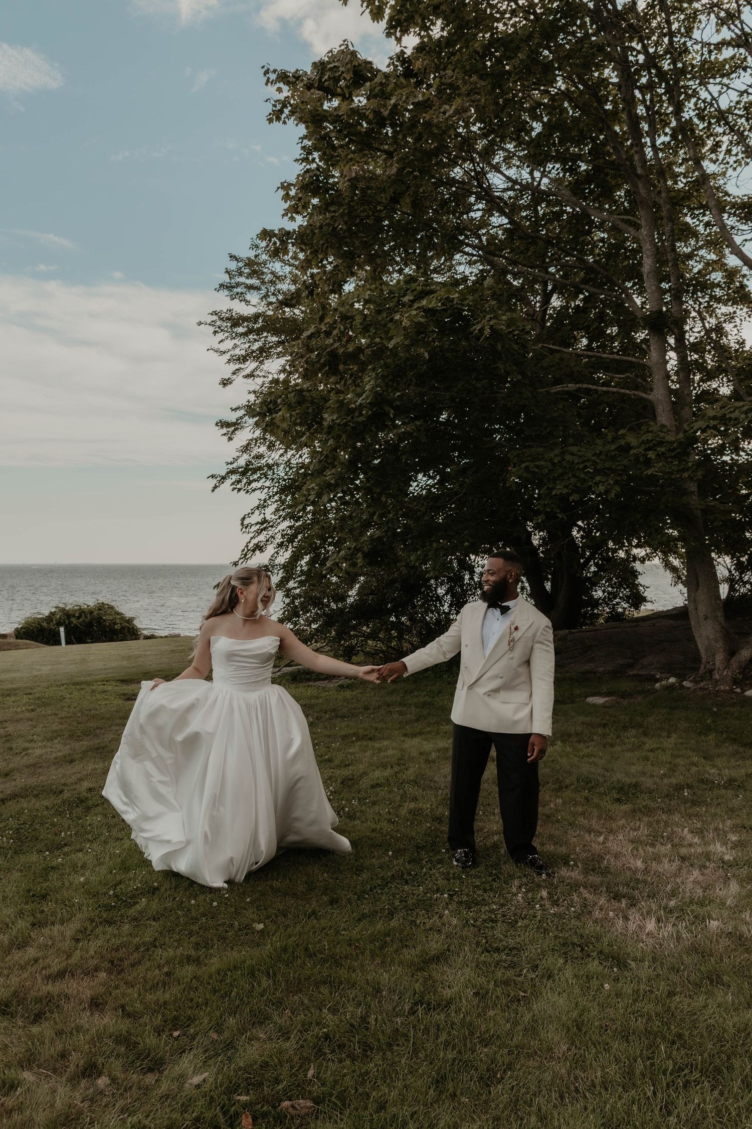 Bride and groom hold hands on green lawn near lake, bride in white gown, groom in white jacket and black pants.