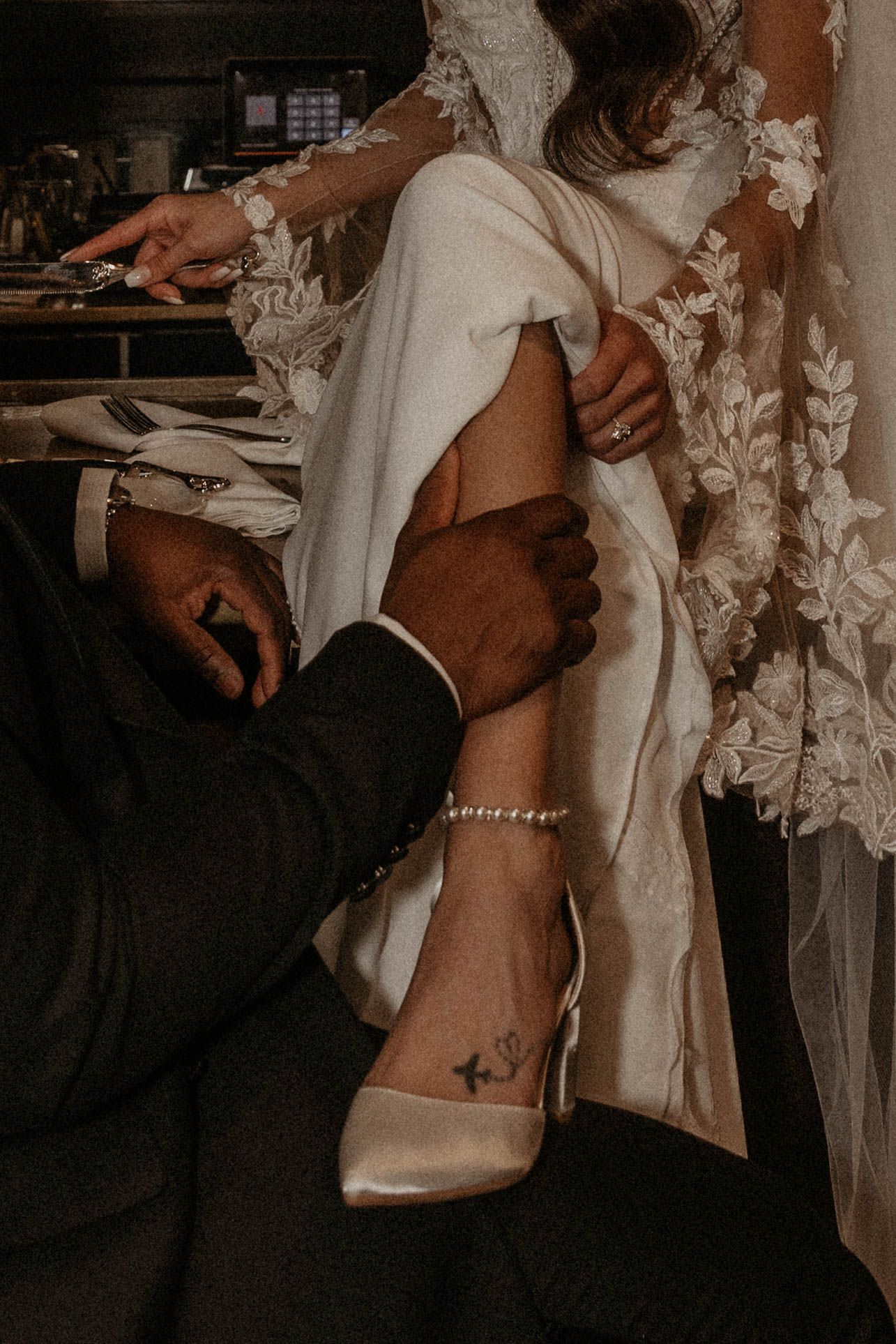 Bride in lace wedding dress with pearl anklet and satin heel, groom in black suit holding her leg during wedding preparation.