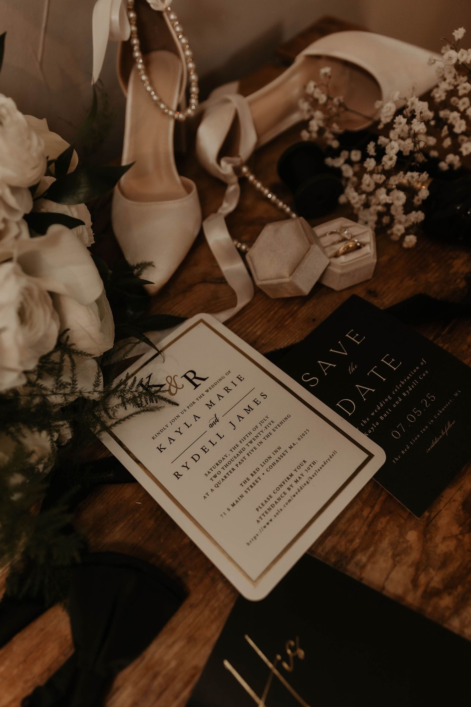Elegant wedding flat lay with ivory heels, floral bouquet, ring box, and monogrammed invitations on a wooden surface.