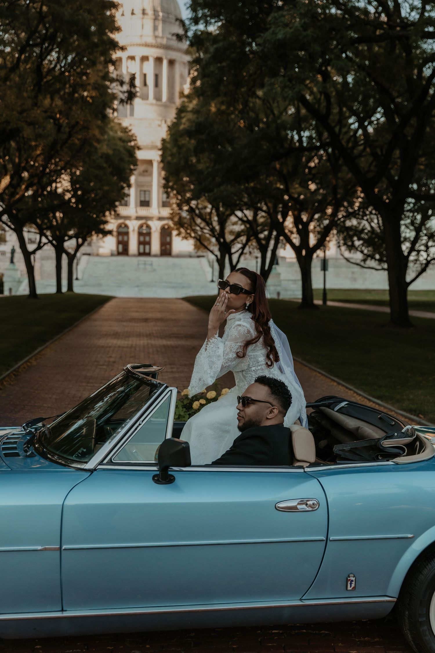 Bride in a white gown and sunglasses sits atop a vintage light blue convertible beside her groom, with a domed capitol building behind.