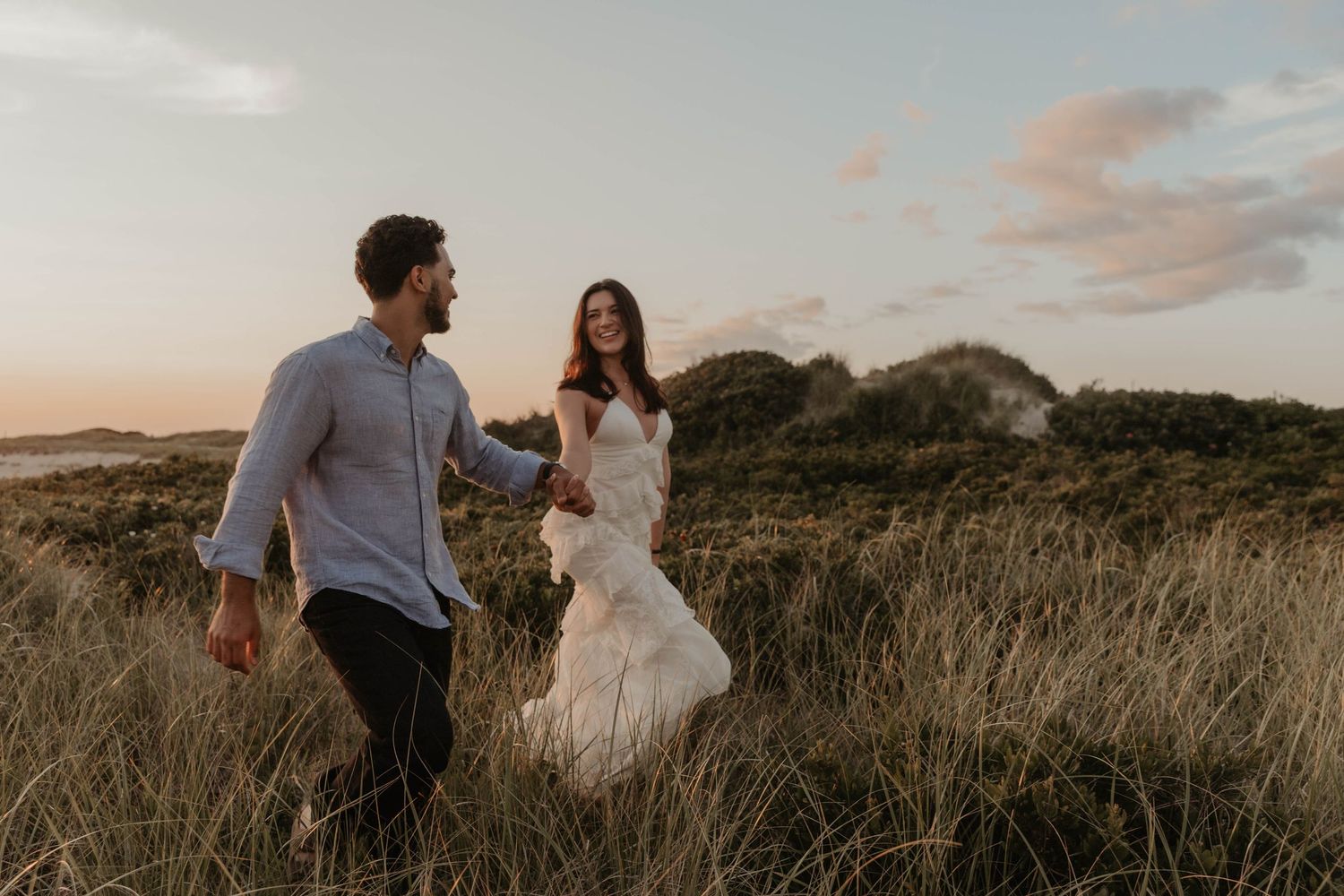 Couple walking hand in hand through golden grass dunes at sunset, bride in white dress laughing joyfully.