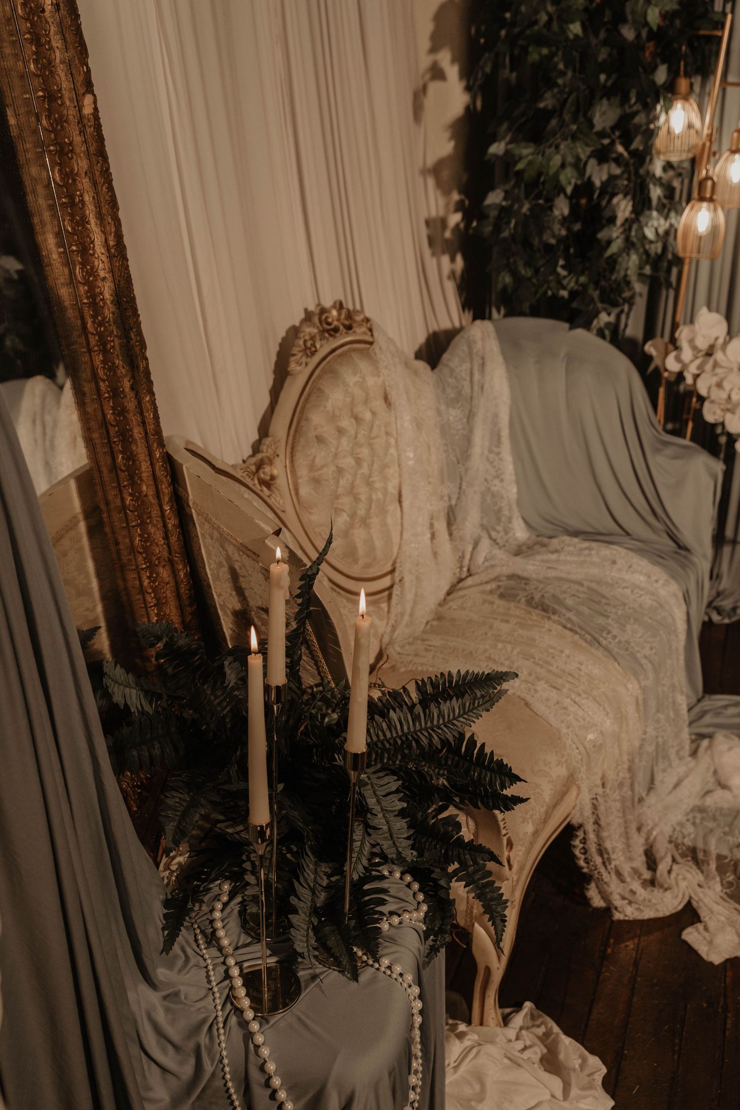 Elegant vintage chair with fur throw, lit taper candles, ferns, and pearl beads in moody styled decor.