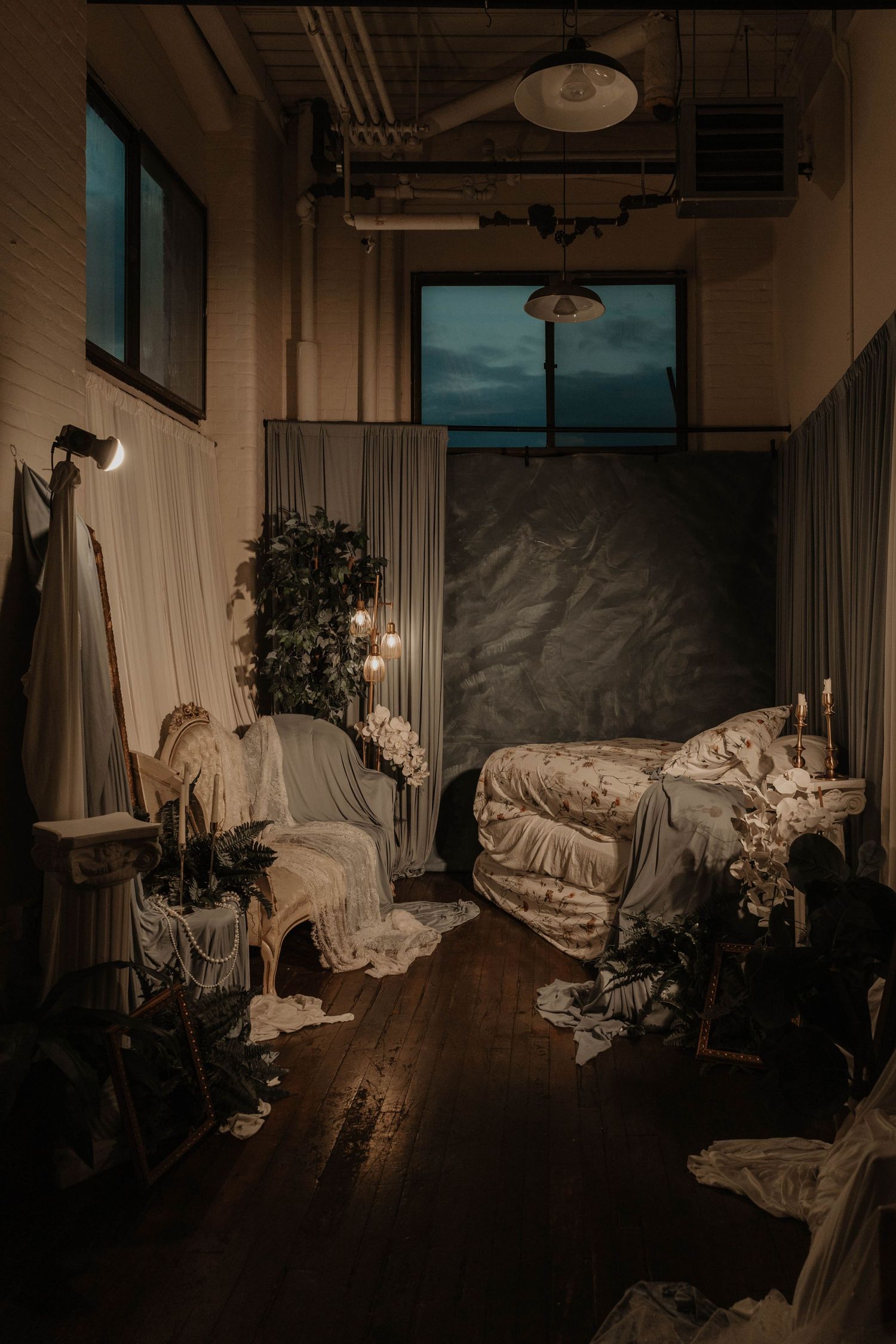 Moody dimly lit bedroom with unmade bed, fairy lights, plants, and draped fabric in a rustic loft space.