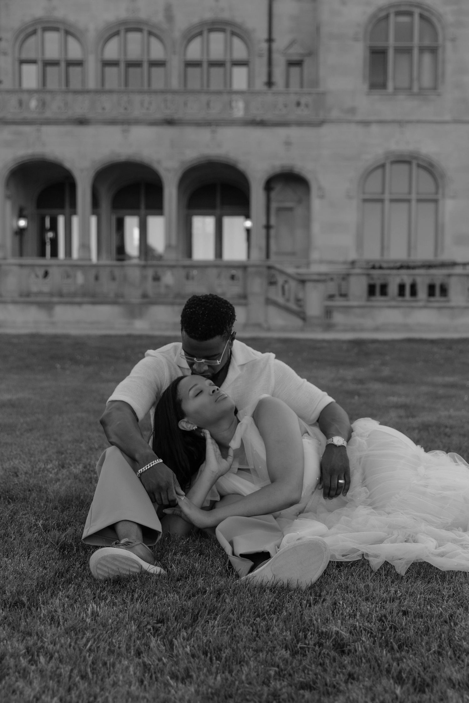 Black and white photo of a couple sitting on grass, bride leaning on groom, with a grand historic building behind them.