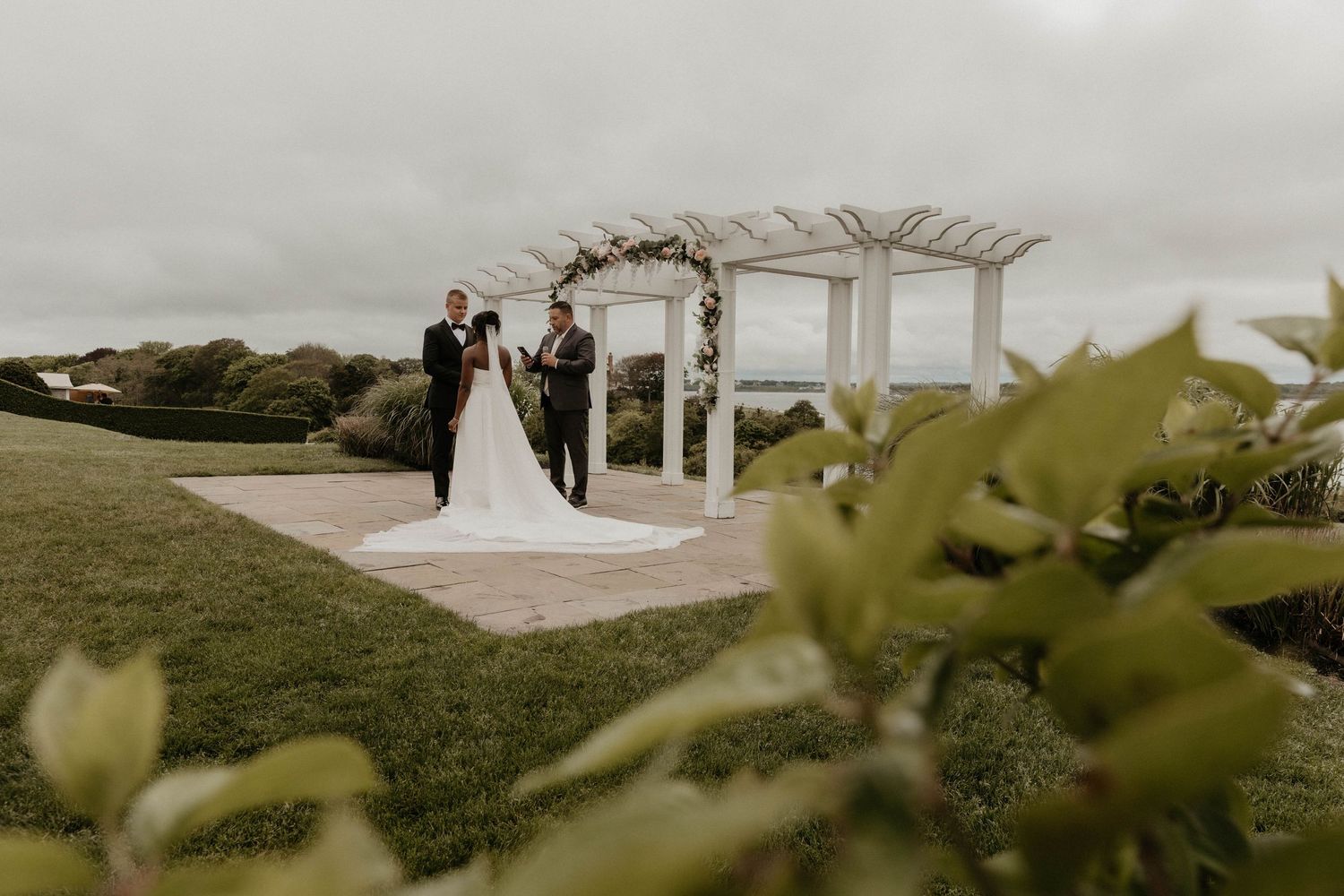 Bride and groom standing under a floral arch pergola on a lush green lawn on a cloudy day.