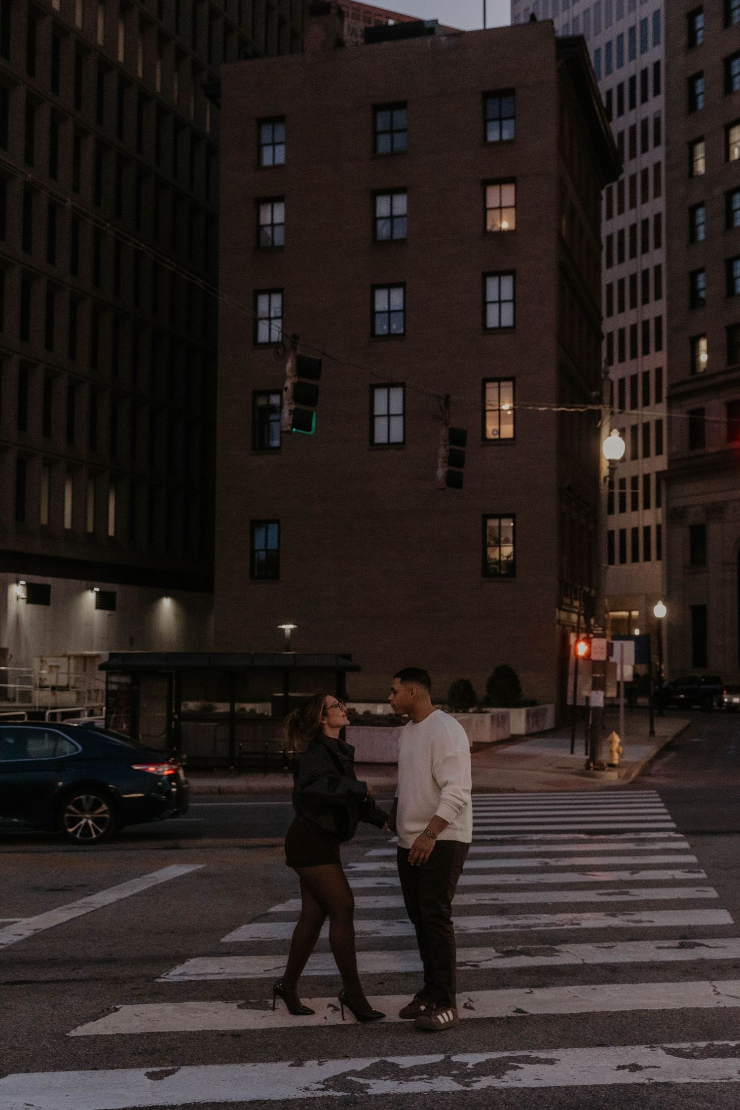 Couple standing on a crosswalk at night in an urban city setting with brick buildings and street lights.