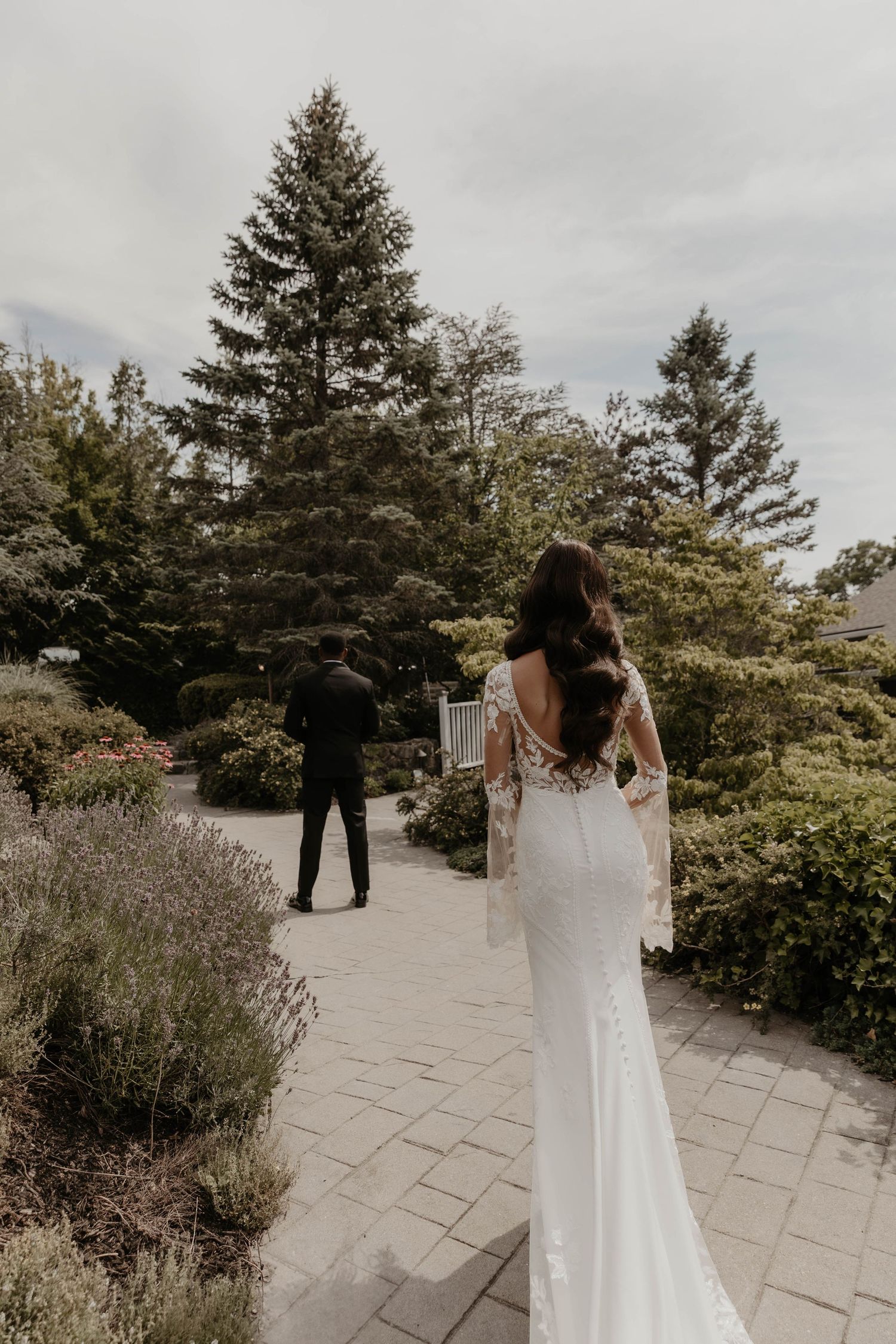 Bride in lace gown walks toward groom on garden path, back detail visible, surrounded by lush greenery.