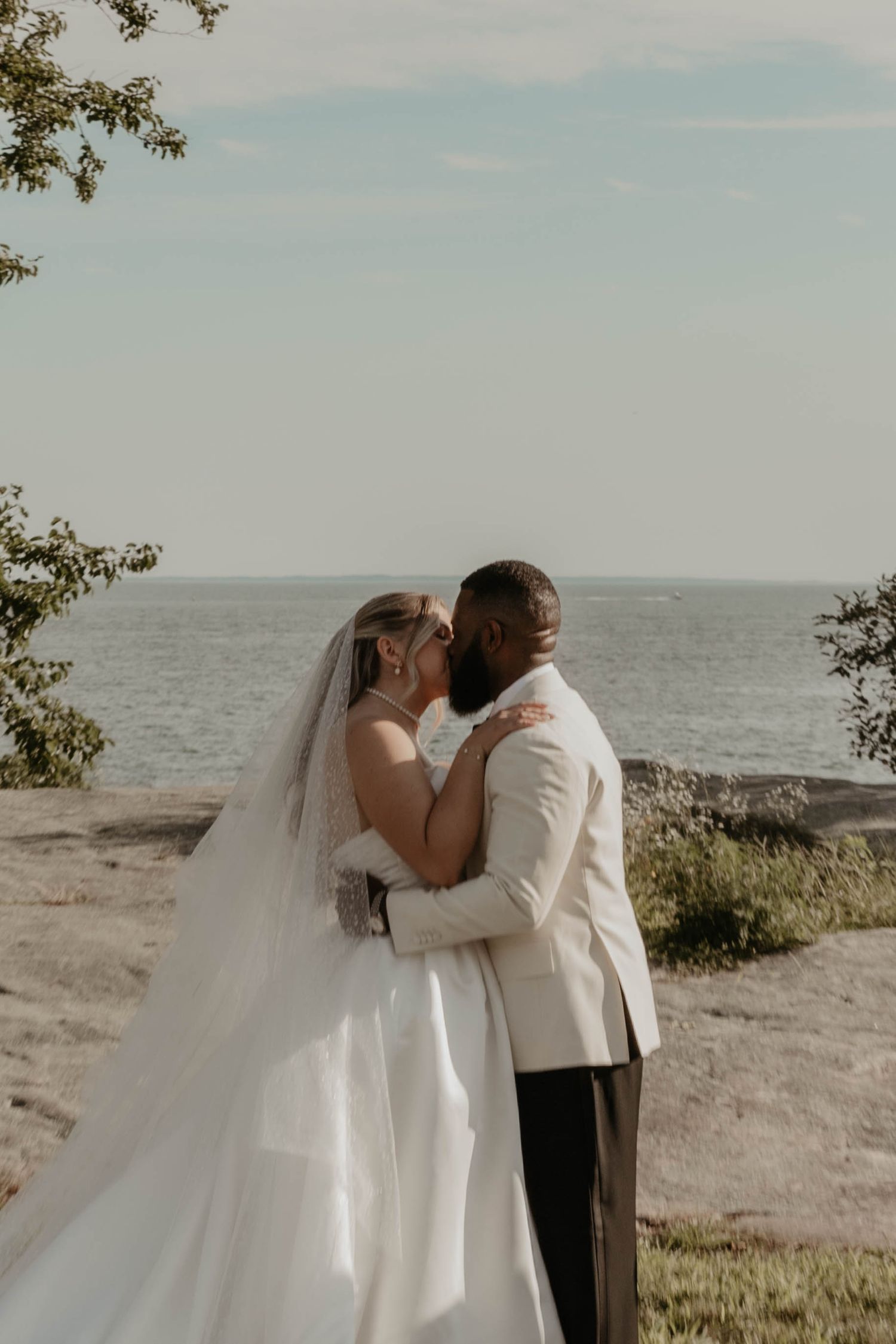 A bride and groom share a romantic kiss by the waterfront, with the bride in a white gown and veil.