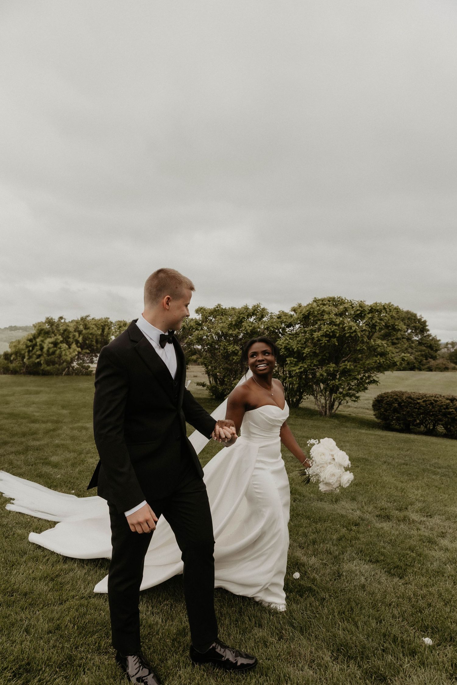 Smiling bride in a strapless white gown holds a bouquet and the groom's hand while walking across a grassy lawn on a cloudy wedding day.