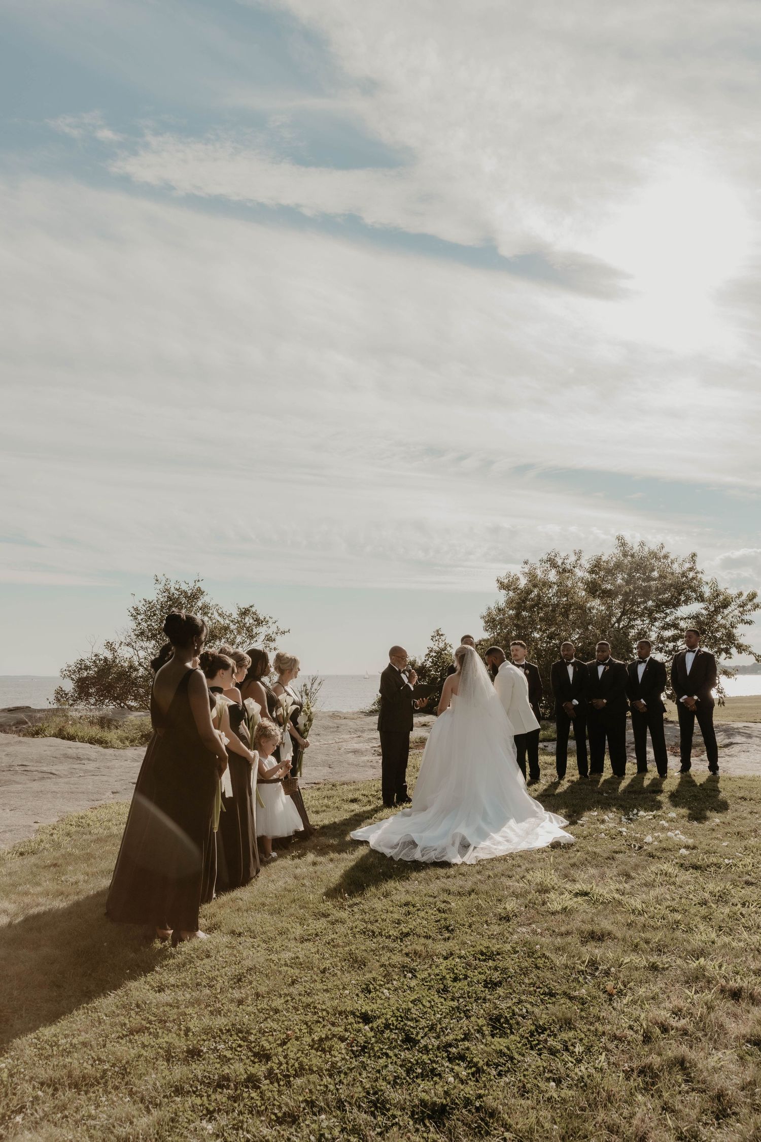Outdoor wedding ceremony on a grassy hilltop with bride, groom, wedding party, and officiant under a cloudy sky.