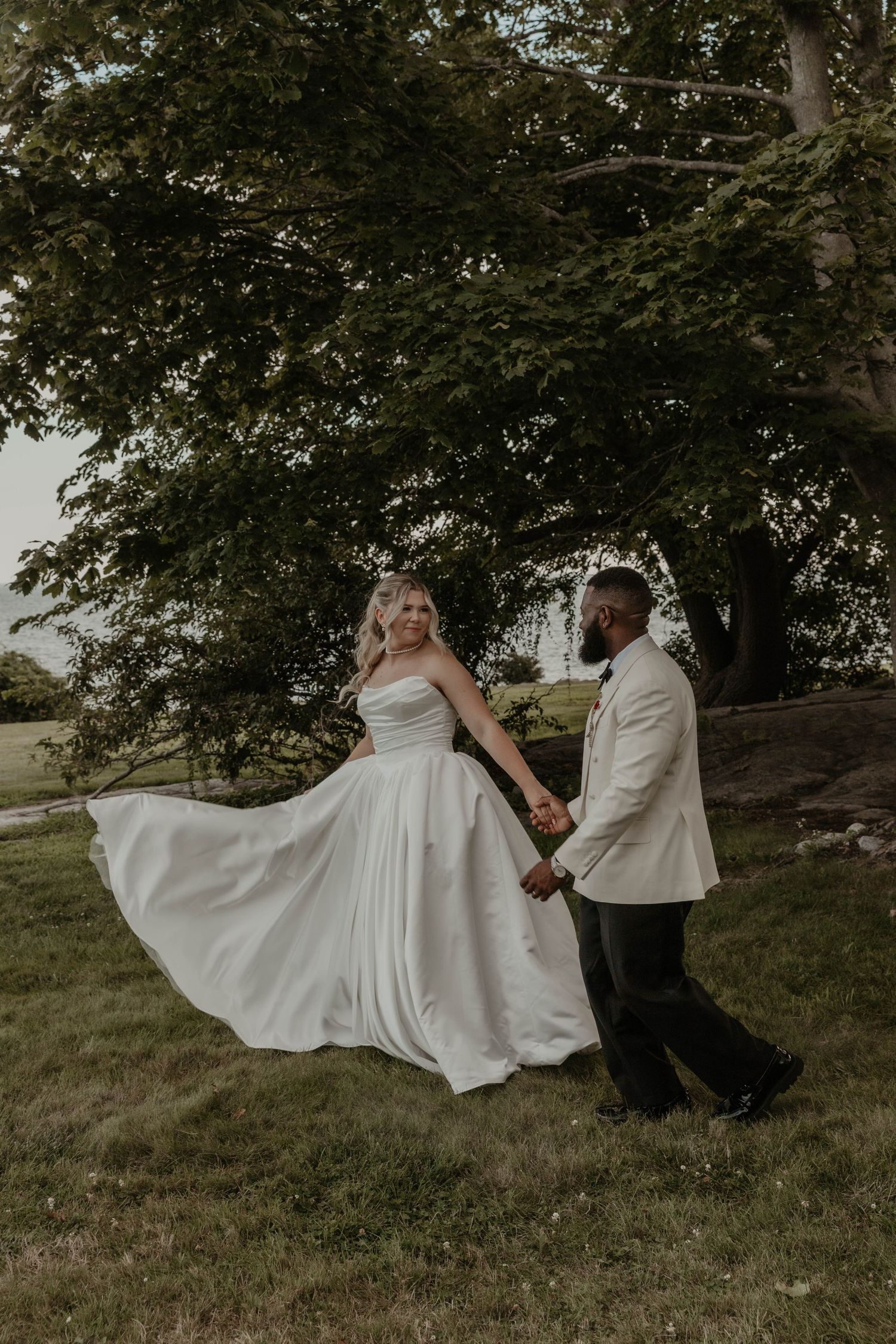 Bride and groom hold hands walking on grass beneath a large tree at outdoor wedding.