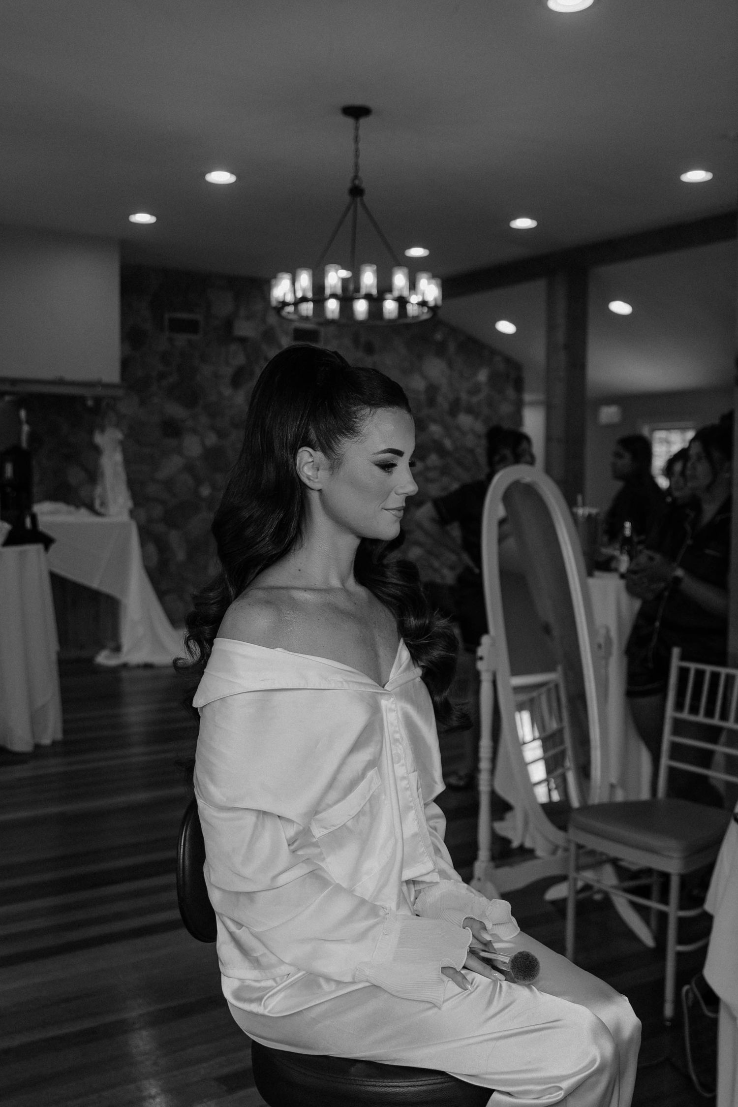 Black and white photo of an elegant bride in off-shoulder white gown seated in a beautifully lit wedding venue.