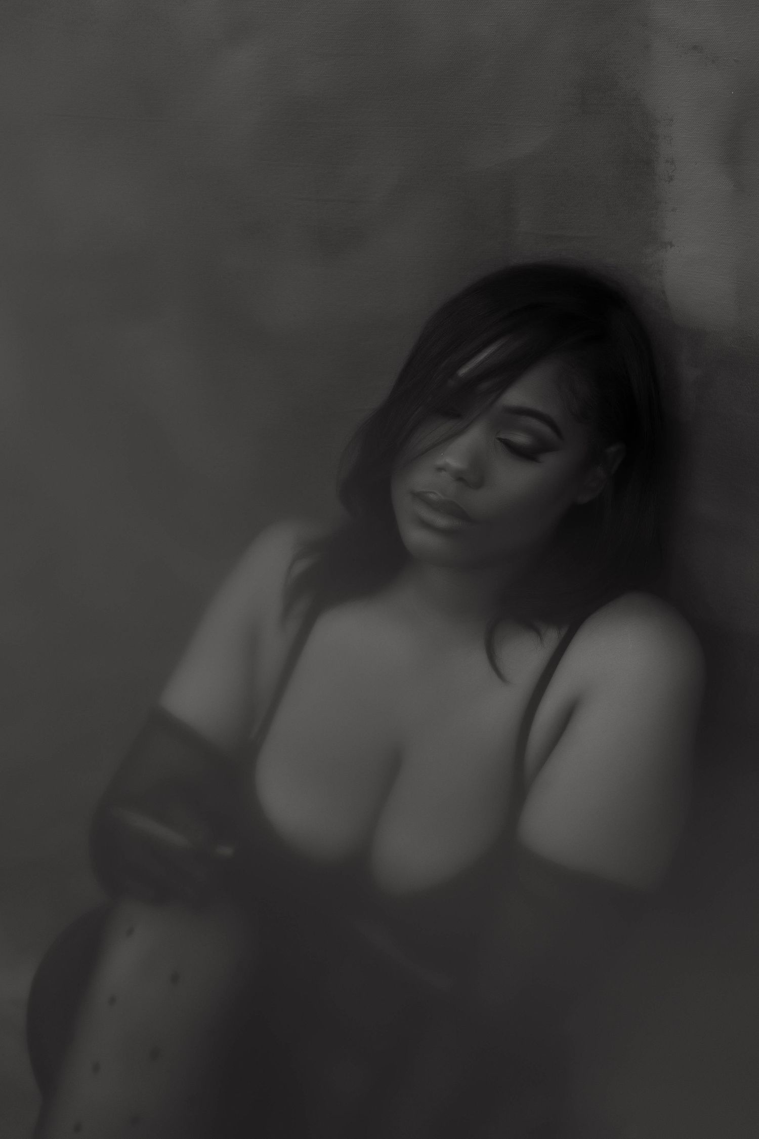 Black and white boudoir photo of a woman in lingerie leaning against a wall in soft focus lighting.