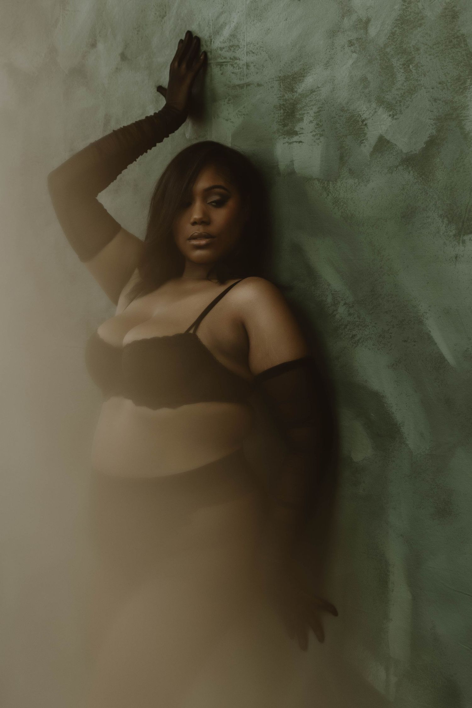 A plus-size Black woman in black lingerie poses confidently against a textured green wall in moody, atmospheric lighting.