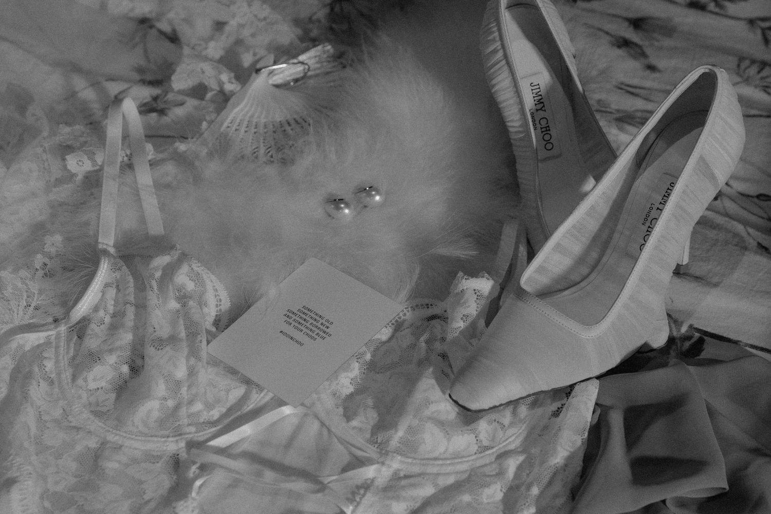 Black and white photo of a high-heeled shoe, feather boa, earrings and a small card resting on crumpled satin fabric.