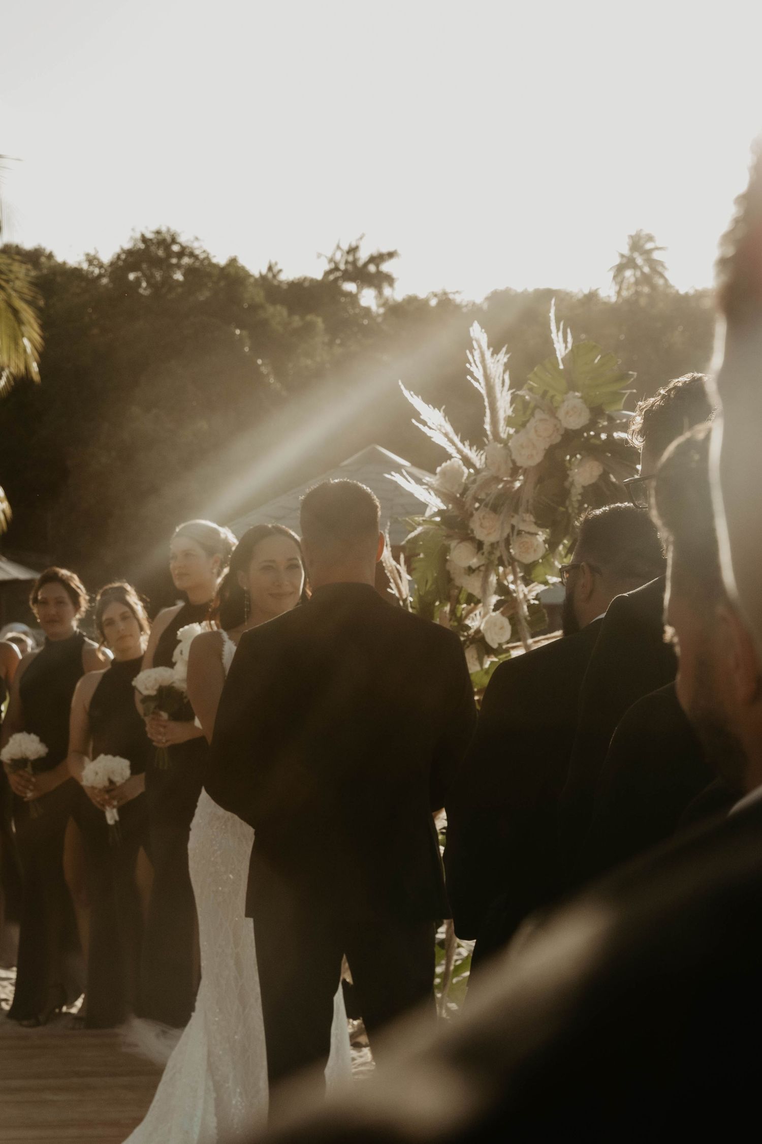 Outdoor wedding ceremony at golden hour with bride and groom at the altar, floral arch, and bridesmaids in black dresses.