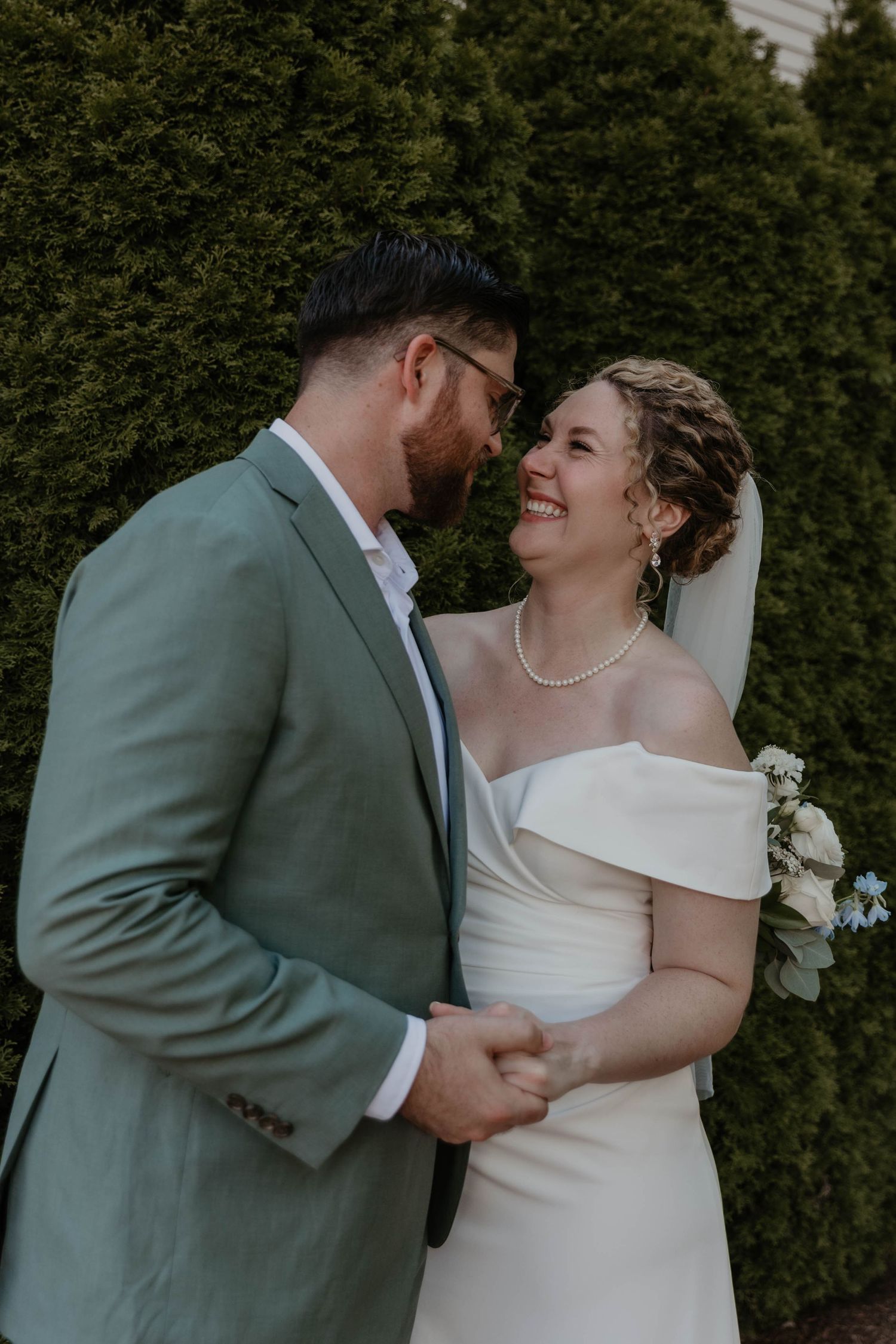 A smiling bride in a white off-shoulder gown holds hands with a groom in a sage suit outdoors.