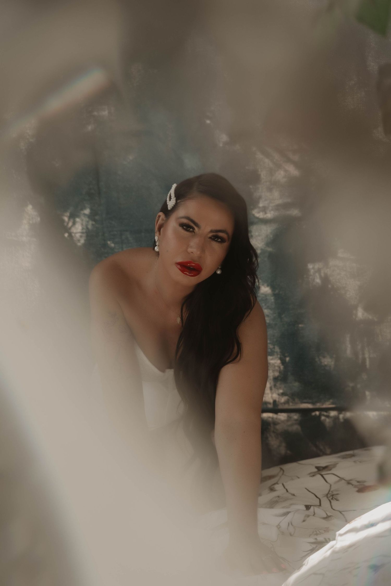 A bride with red lips, dark hair, and a white hair accessory gazes through blurred foliage in a moody portrait.