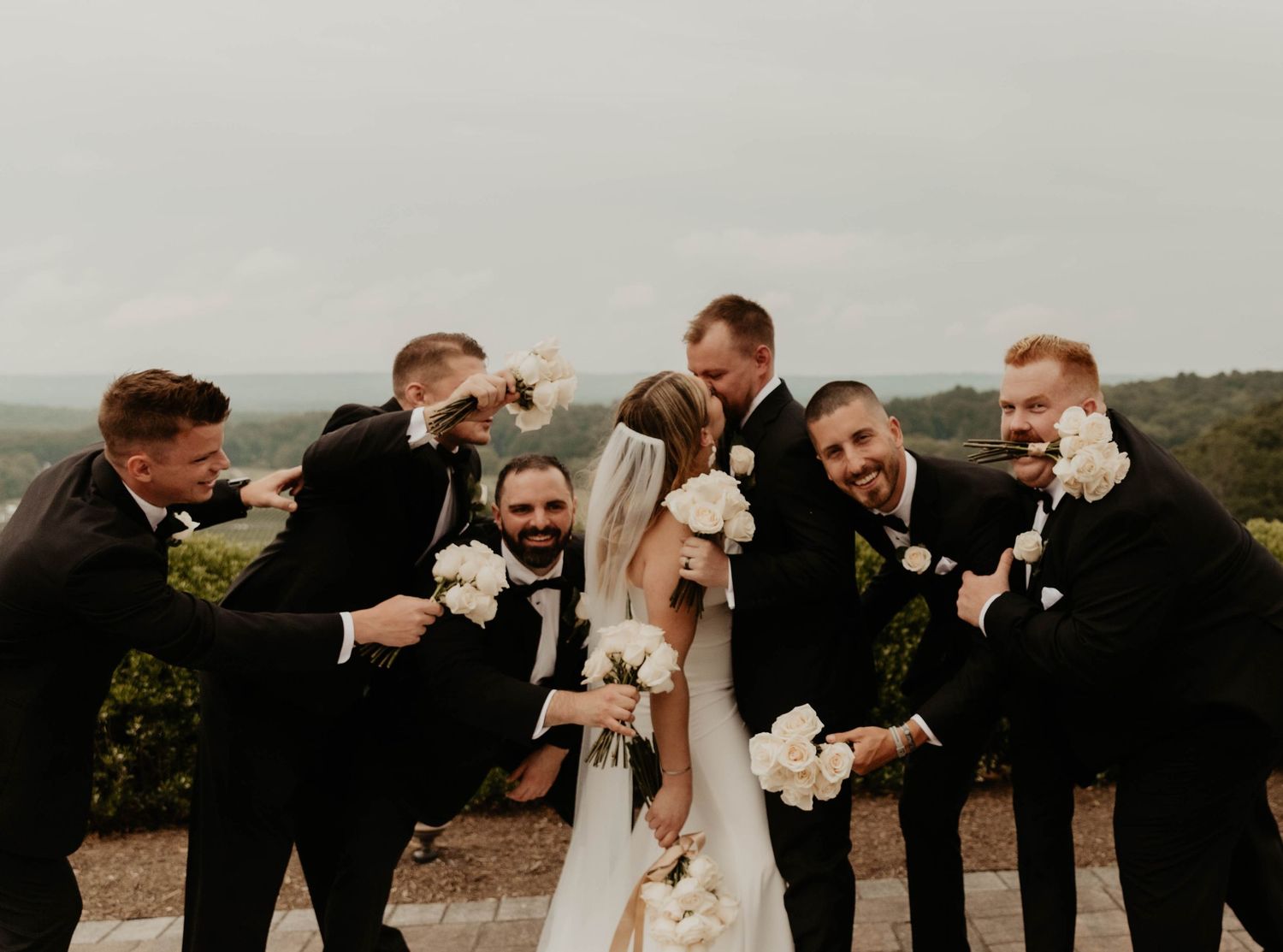 Bride in a white gown and veil stands among six groomsmen in black suits as they playfully lean in with white floral bouquets outdoors.