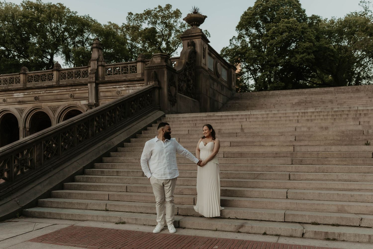 Couple holding hands on stone steps at Bethesda Terrace in Central Park, bride in white gown and groom in light suit.