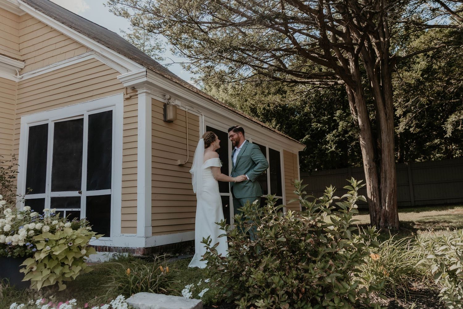 Bride and groom share a tender first look moment outside a charming yellow cottage surrounded by greenery.