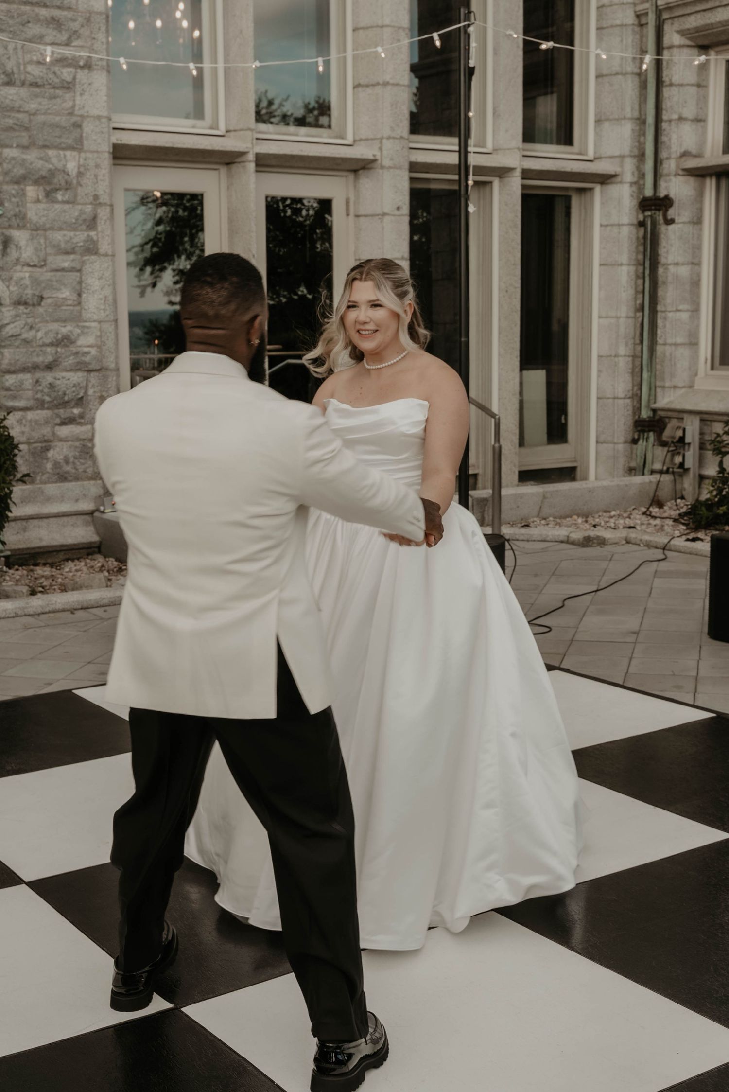Bride and groom share their first dance on a black and white checkered floor outside a stone venue.