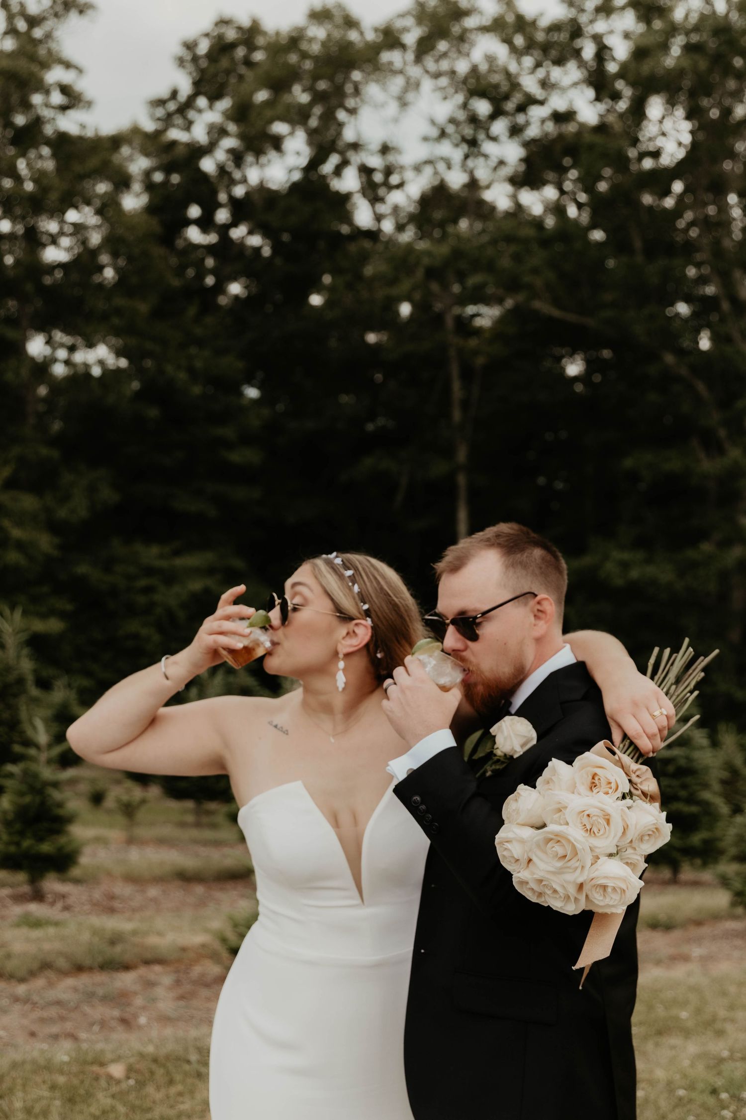 Bride in a white strapless gown and groom in a dark suit and sunglasses drink from glasses while holding a cream rose bouquet outdoors.