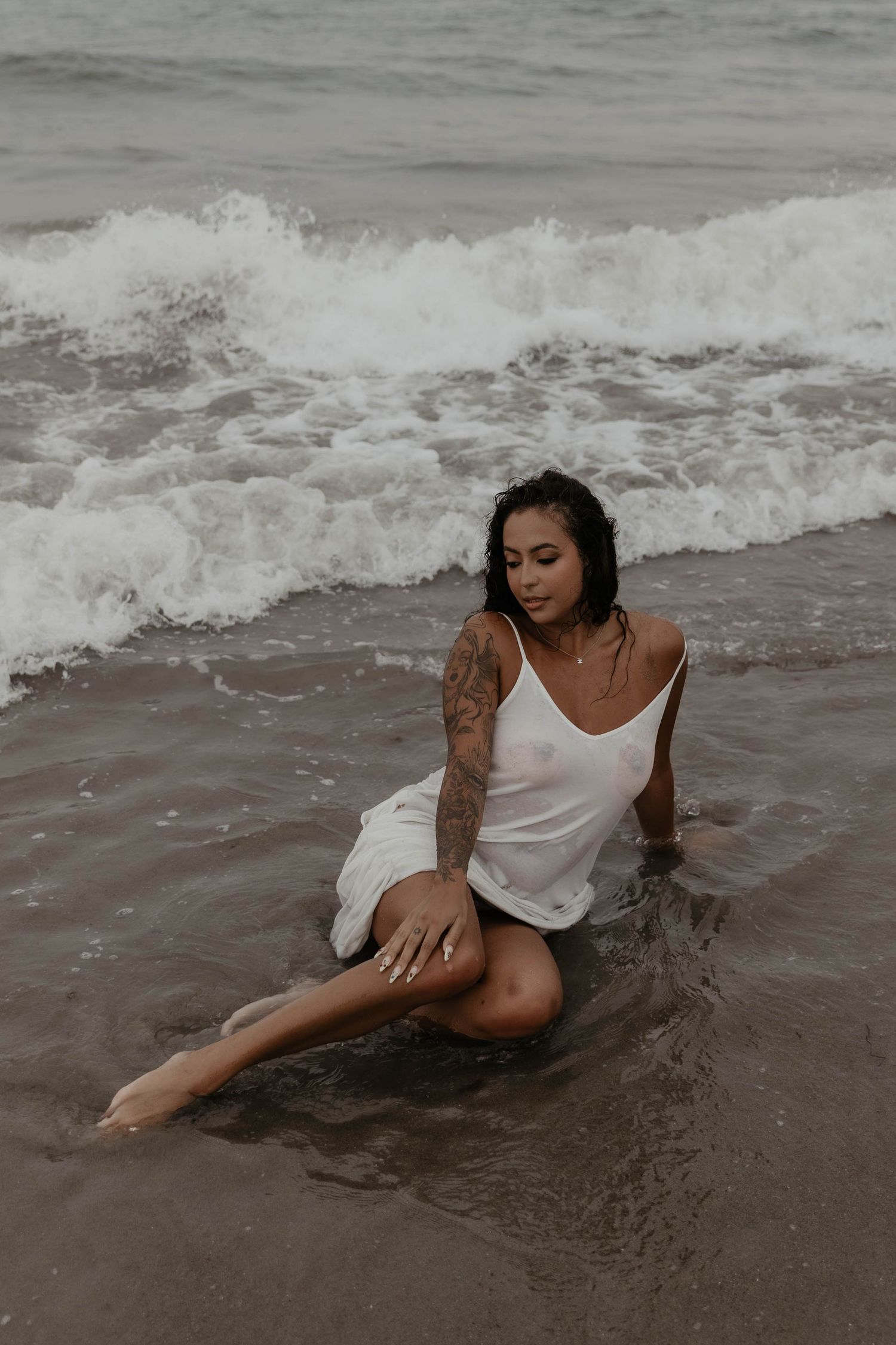 Tattooed woman in white dress sitting in ocean waves on a sandy beach.