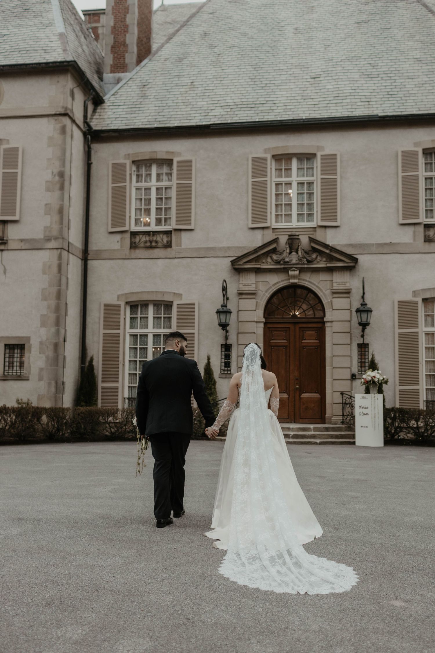 A bride in a white lace gown and veil walks hand-in-hand with a groom toward an elegant stone manor.