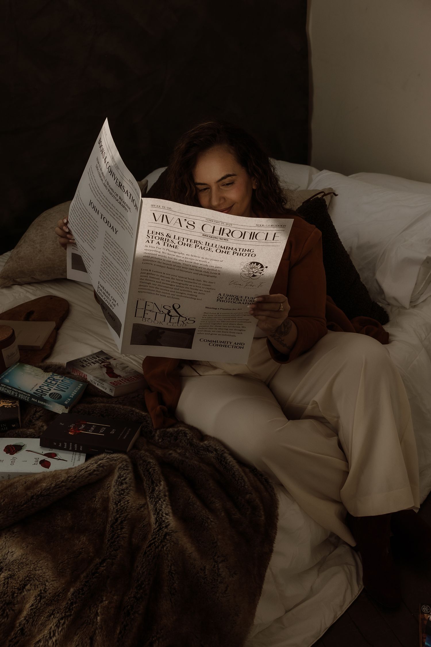 Woman reading Viva's Chronicle newspaper while relaxing in bed surrounded by books and cozy blankets.