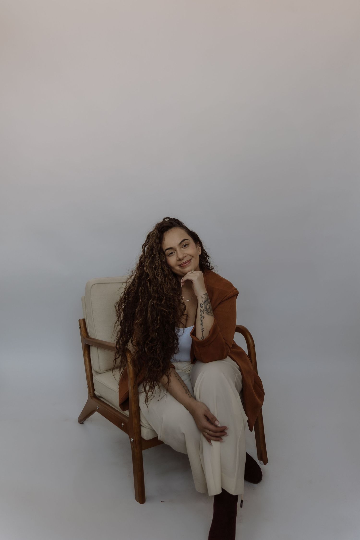 Woman with long curly hair sits smiling in a wooden chair, wearing a brown jacket and white pants.