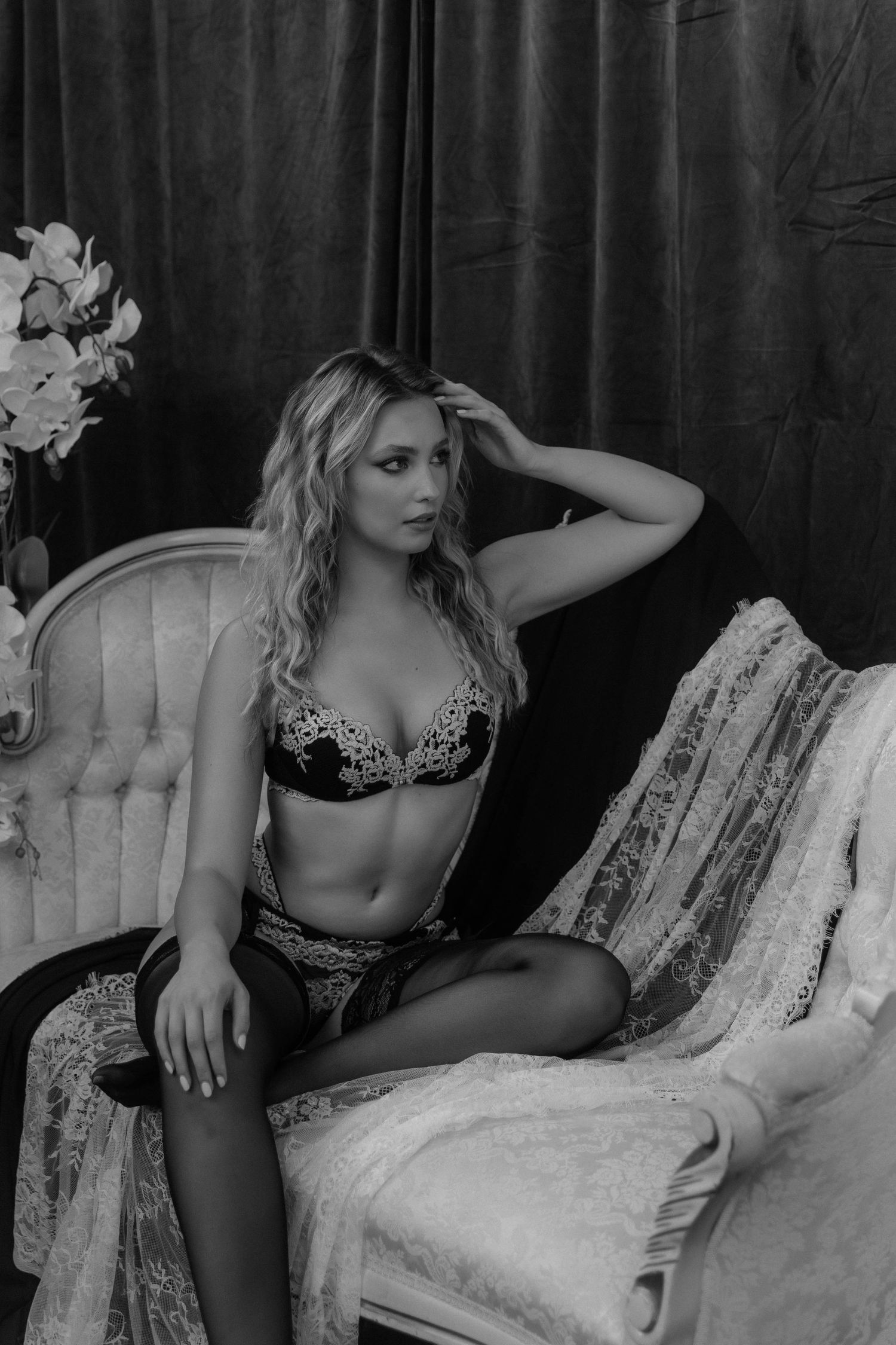 Black and white boudoir photo of a blonde woman posing elegantly in lace lingerie on a vintage chair.