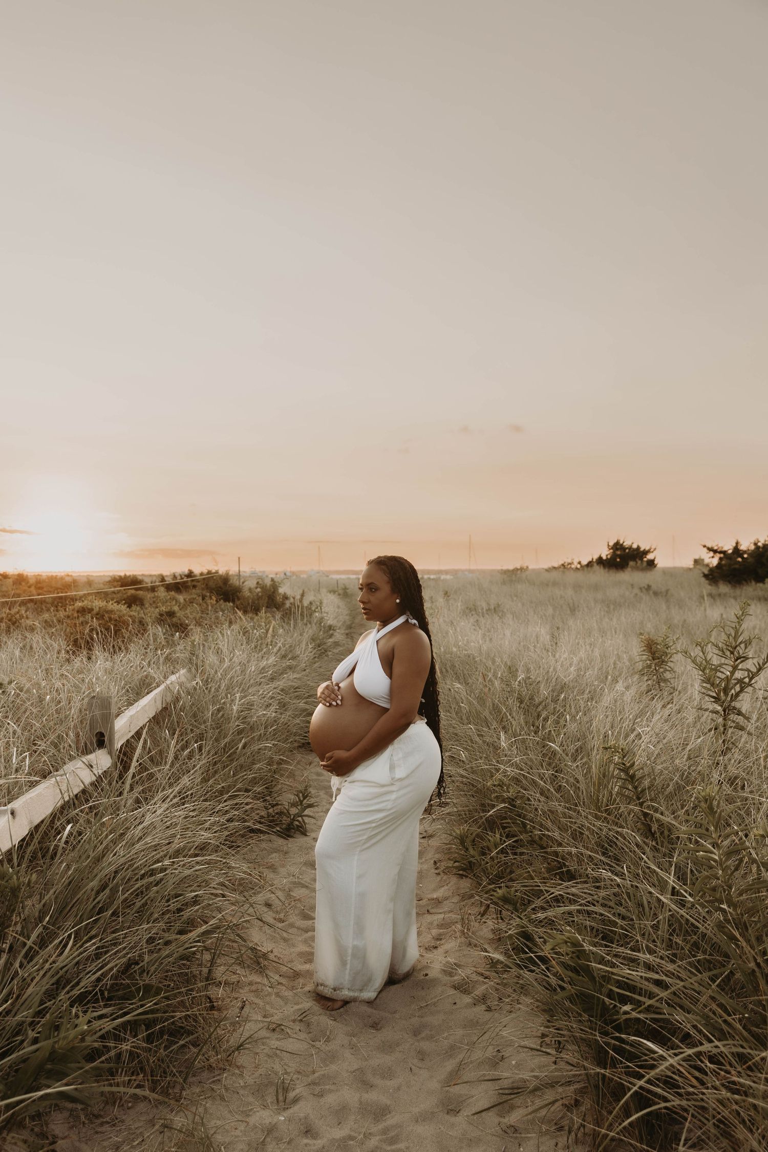 Pregnant woman in white dress cradling baby bump on sandy beach path at sunset.